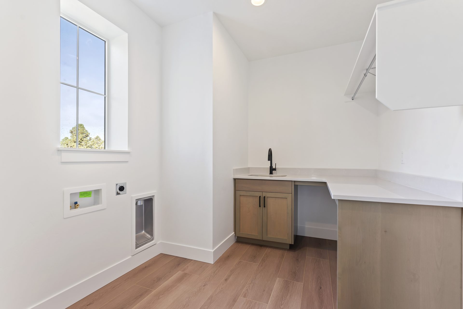 A laundry room with an L shape vanity to the right with wood color base and light color countertops, light color walls, brown wood texture flooring and a window.
