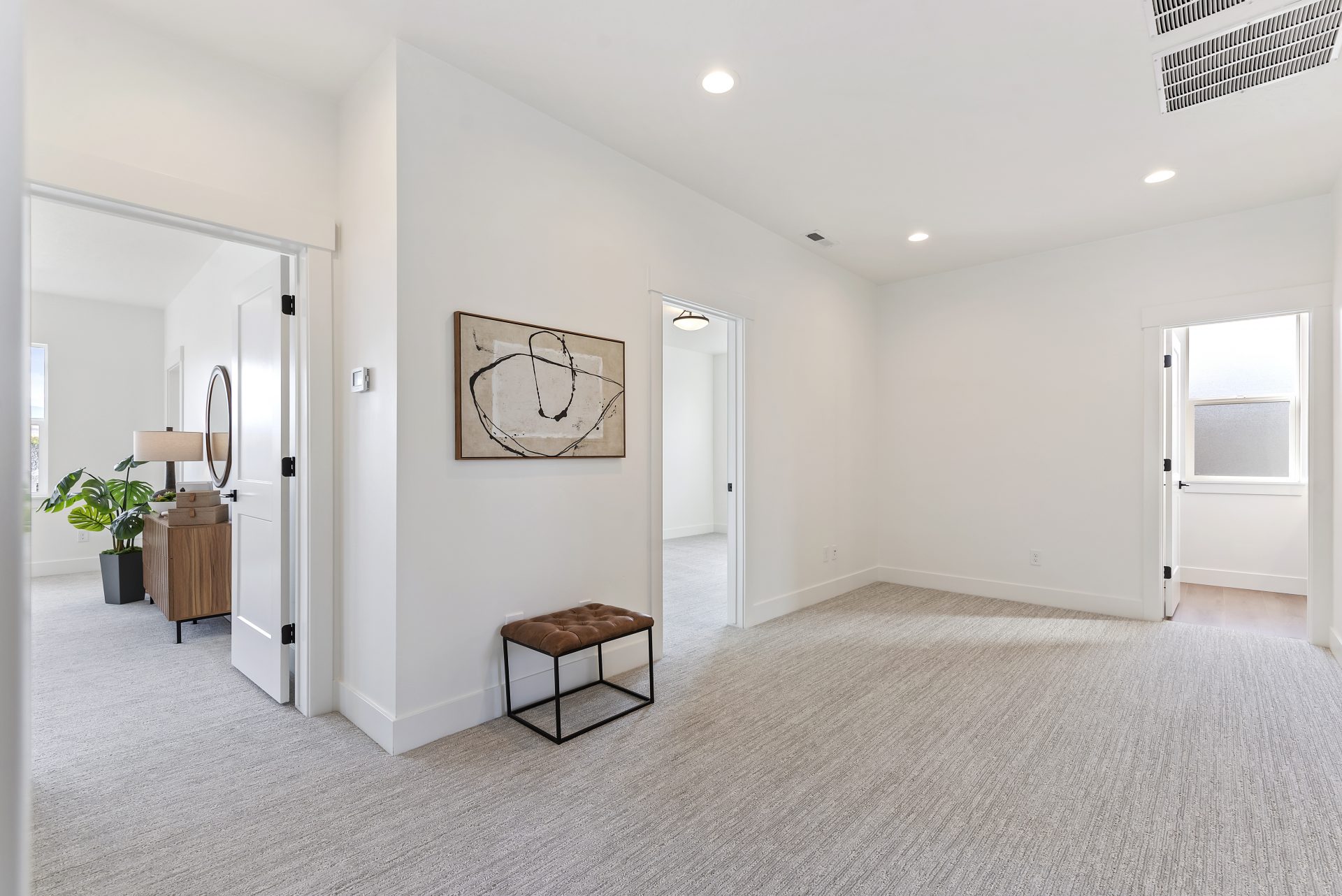 A large hallway with three open doorways, light color carpet, light color walls a hanging piece of art on one wall with a small bench underneath. In the distance through a doorway to the left, you can see bedroom furnishings.