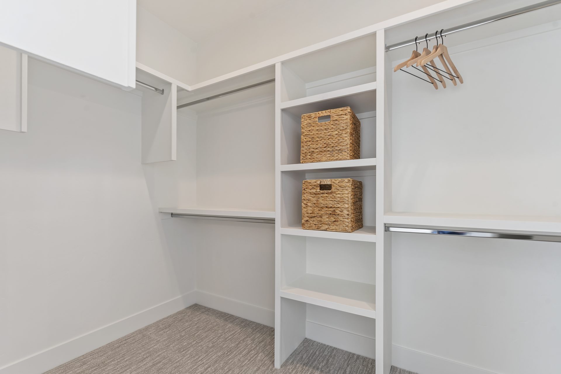 A bedroom closet with multiple shelves and metal hanging rods, with 2 baskets and 4 hangers hanging with gray carpet flooring.