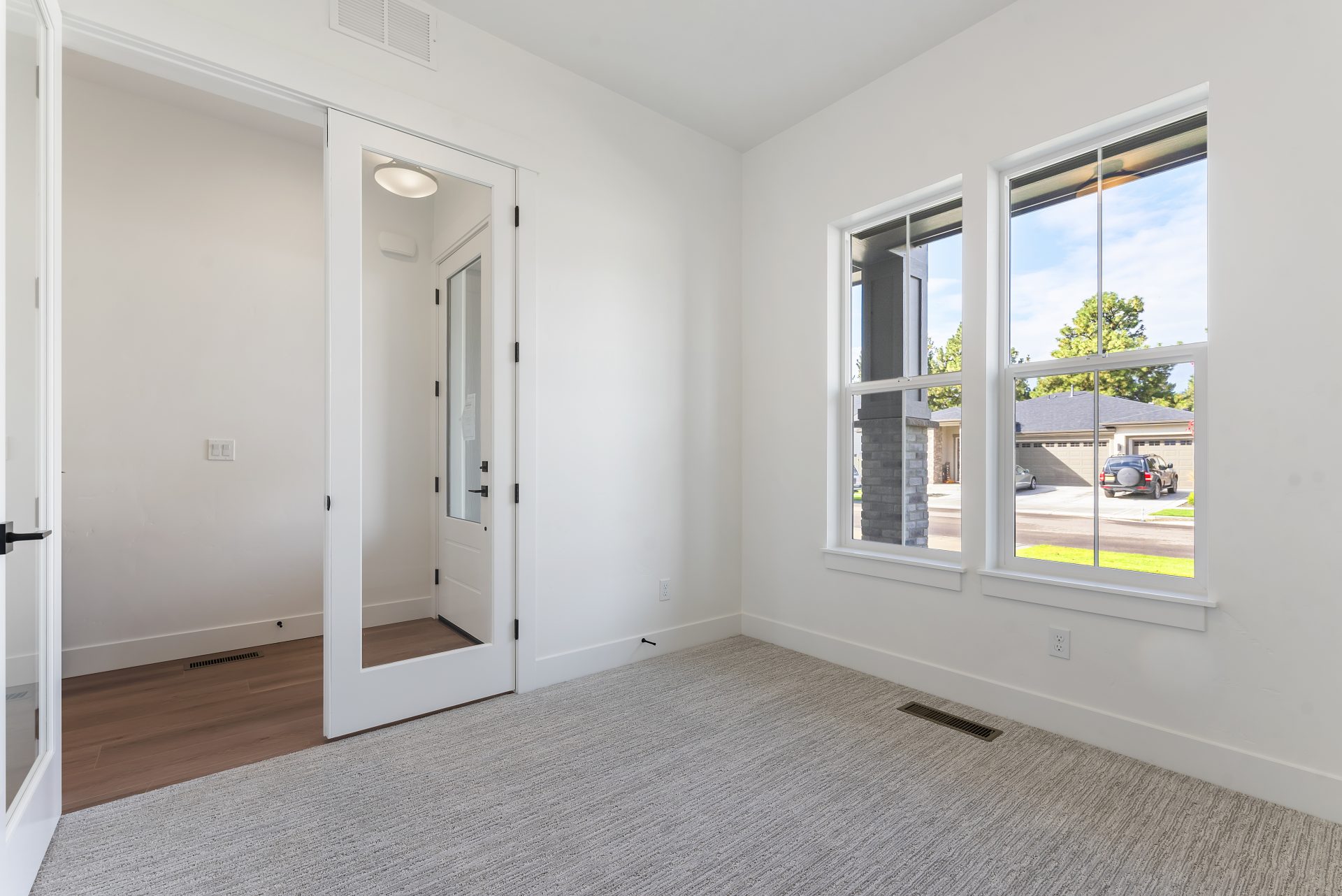 A room with gray carpe flooring, two windows, light colored walls, and a double glass pane entry door with one door open.