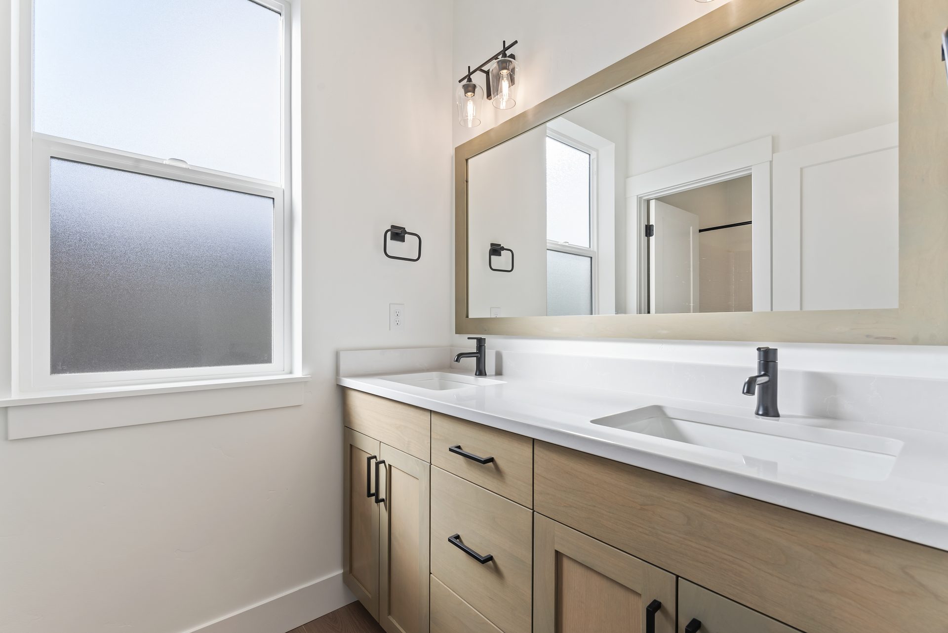 View of a bathroom with a double vanity sink, with medium brown stained wood base, white countertops and short backsplash with black fixtures, and a mirror spanning the length of the vanity with a matching wood trim around the mirror. Above the mirror is 