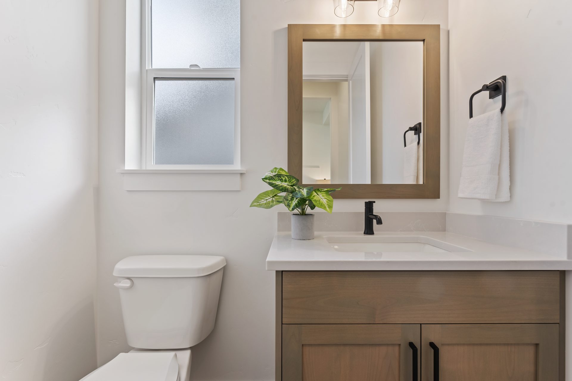 View of a powder room with with a single sink vanity with brown stain wood base, white countertop, black faucet, with a mirror and matching brown trim around it. To the left of the vanity is a toilet with a fogged glass window above it.