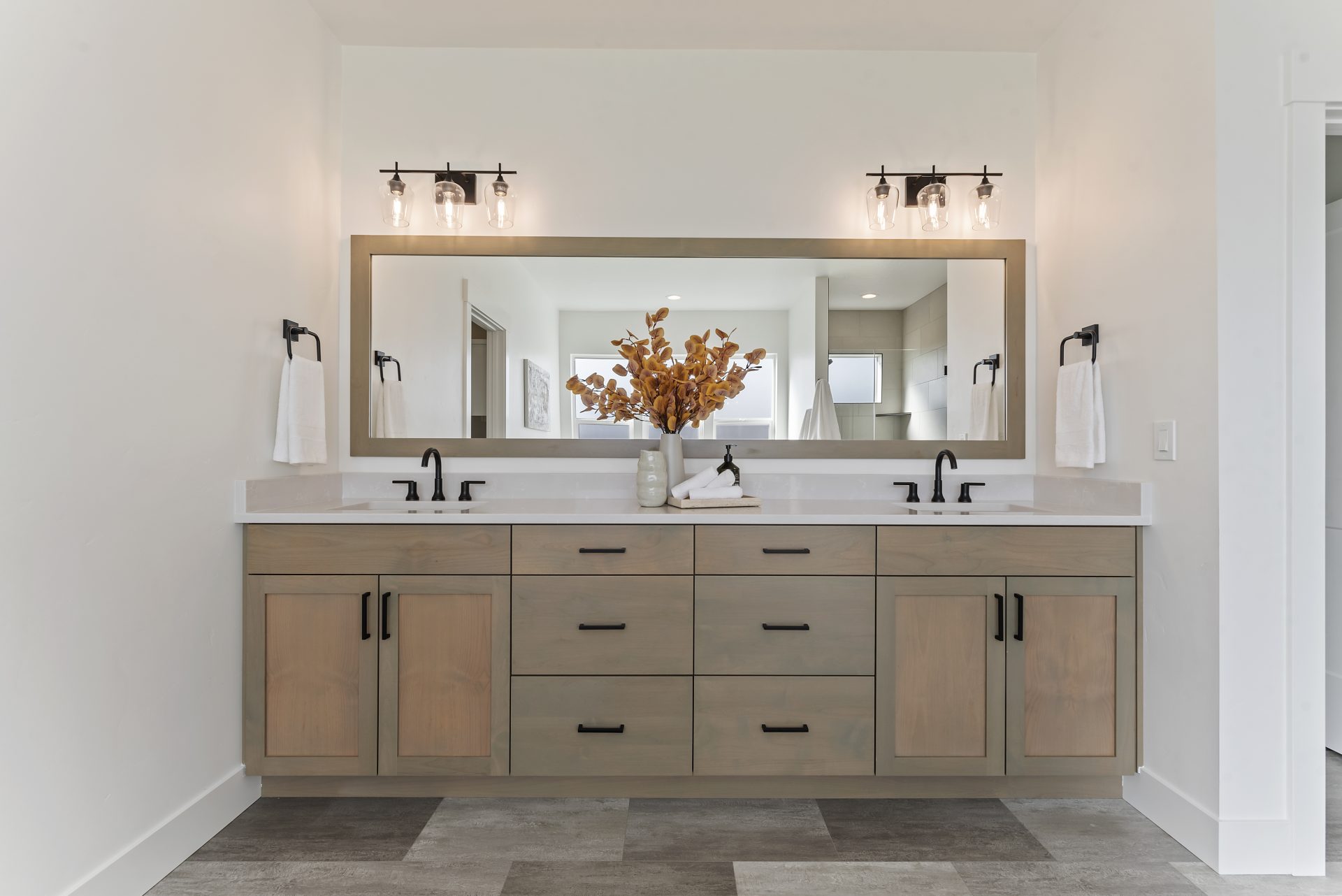 A long bathroom vanity with double sinks, black faucets, brown stained wooden base, a long mirror with matching wood trim around it spanning the length of the vanity with two- 3 light fixtures above the mirror. There are handowel holders with towels on ea