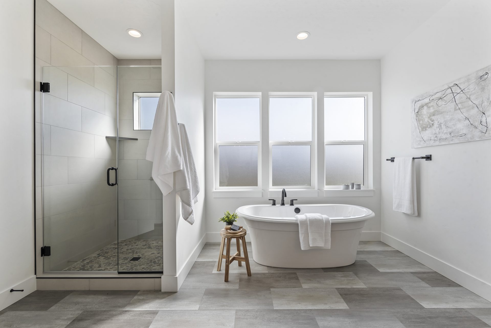 A bathroom with lighter and darker floortiles with a free standing tub with black faucet and handles. Behind the tub are three frosted glass windows, to the right a light colored wall with a black towel hanger with towel and hanging art piece. To the left