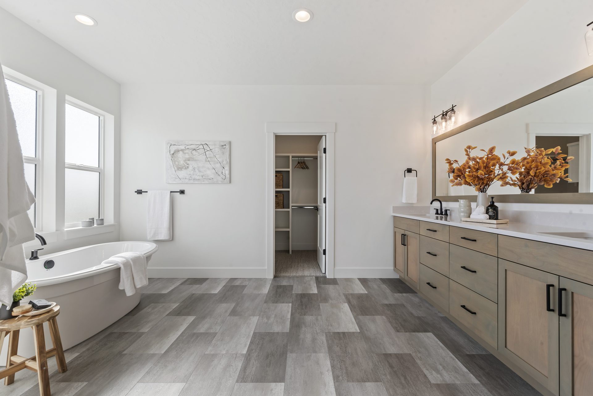 A large bathroom with a light and darker tile floor, to the left a white free standing tub with a black faucet and frosted windows, straight ahead is open open door leading to a walk in closet with shelving. To the right is a long pantry with a stained wo