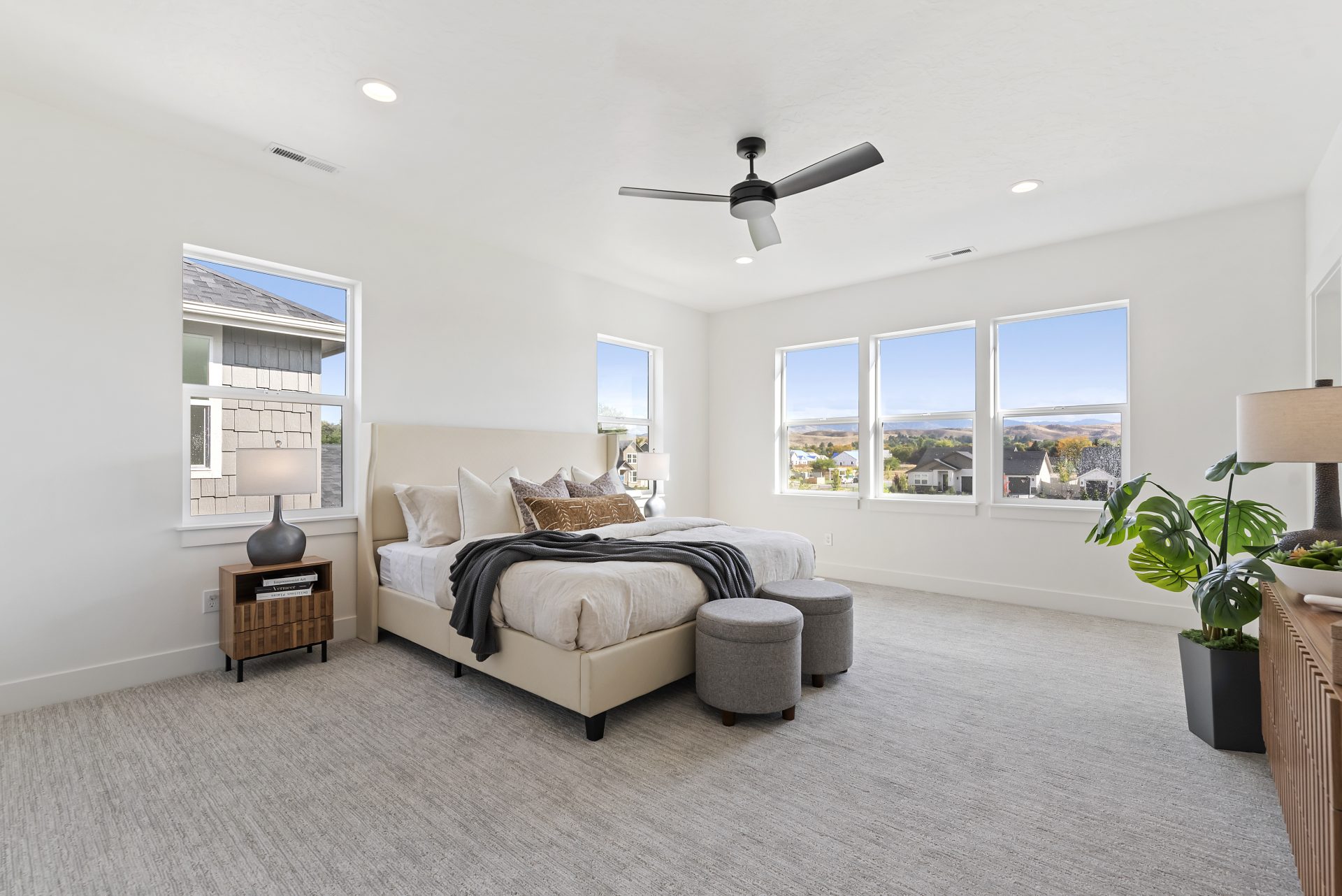 A large bedroom with gray carpet flooring, a bed with a cream color fabric frame, light color walls, a dark color ceiling fan, with 5 windows creating a bright space.