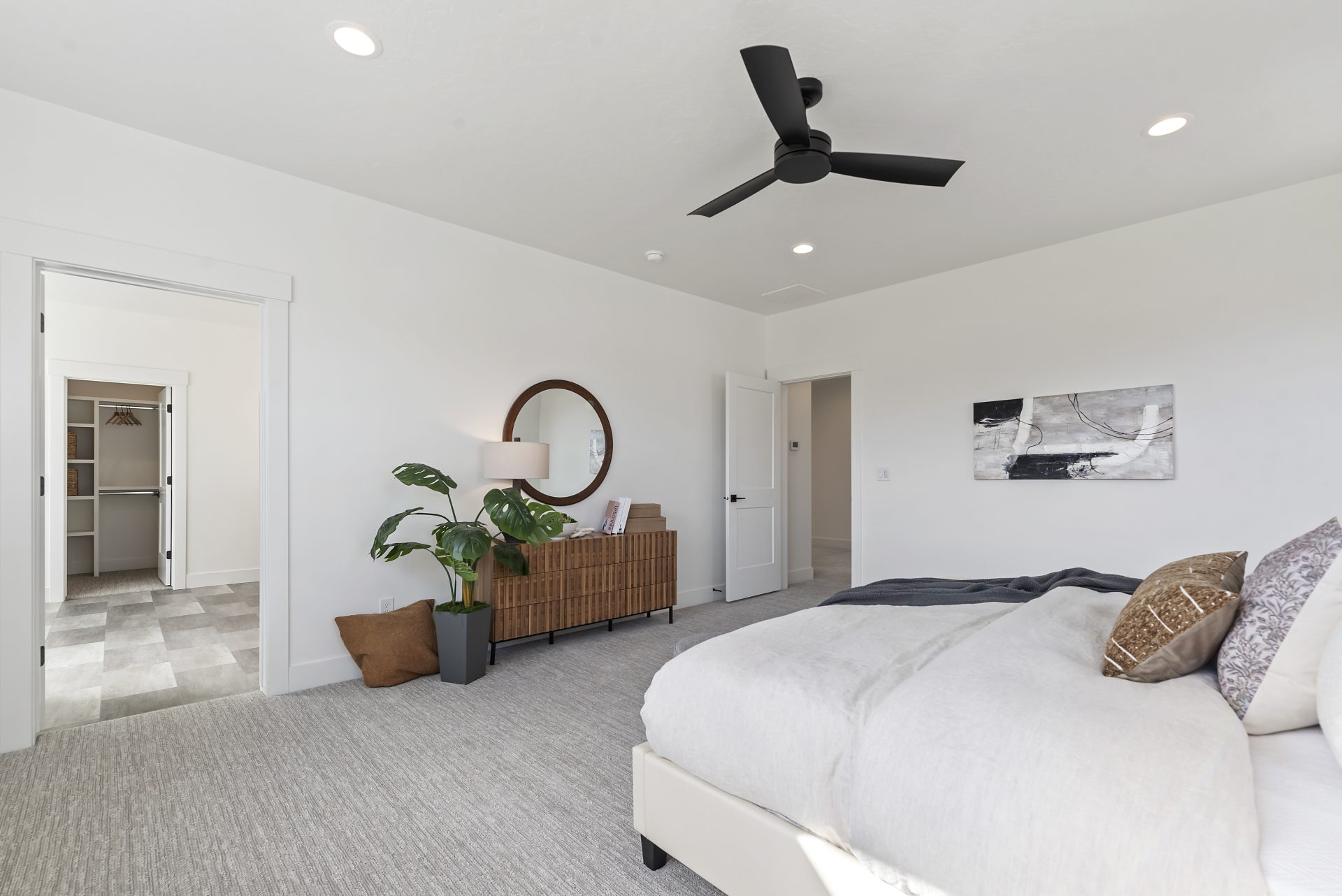 A large bedroom with gray carpet flooring a brown wood stain look dresser with decorations on top, a plant in a black container, to the right an open doorway leading out of the room and on the wall nearby a hanging piece of art. To the left is the doorway