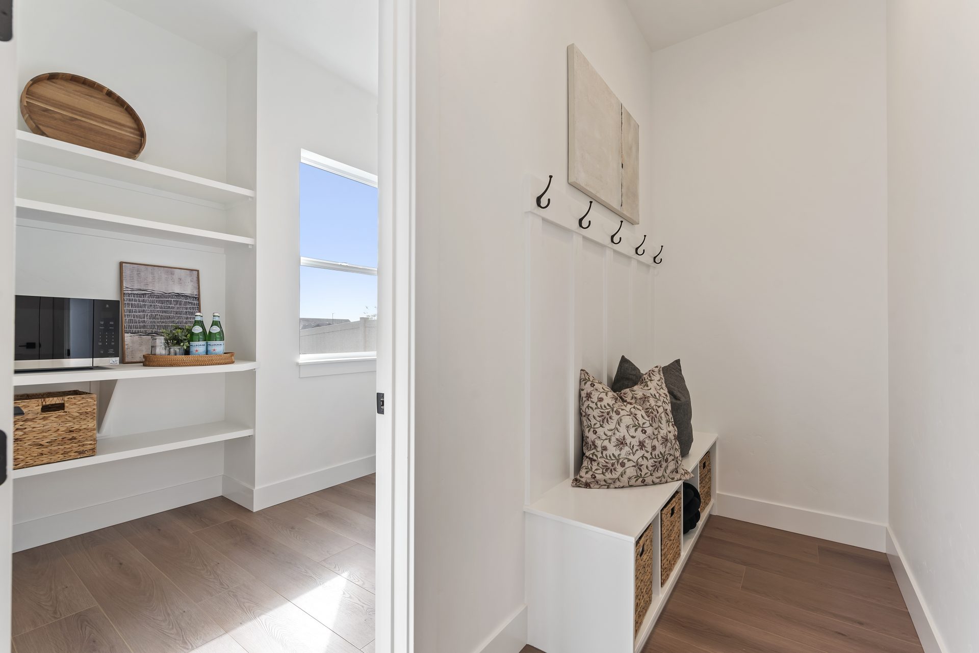 A mud room with a bench with three cubby spaces underneath. Above the bench is a board with 5 coat hooks, light color walls, brown stained wood texture flooring, to the left is an open doorway leading to the panty.