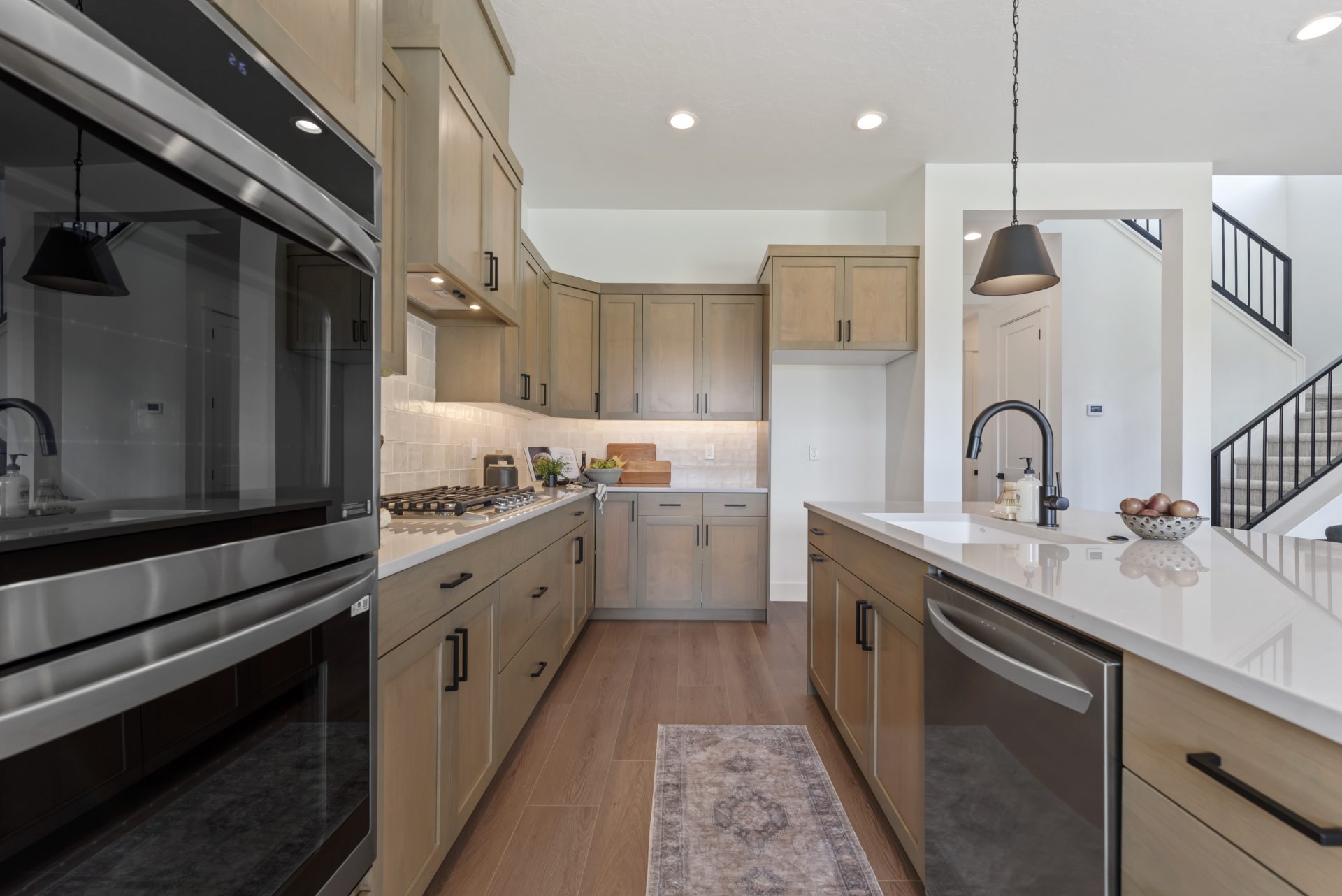 A kitchen view between the kitchen island on the right and upper and lower cabinets on the left. Also on the left are the double ovens, gas range cooktop with light color tile from countertops to cabinets. To the right is the island with a stainless steel