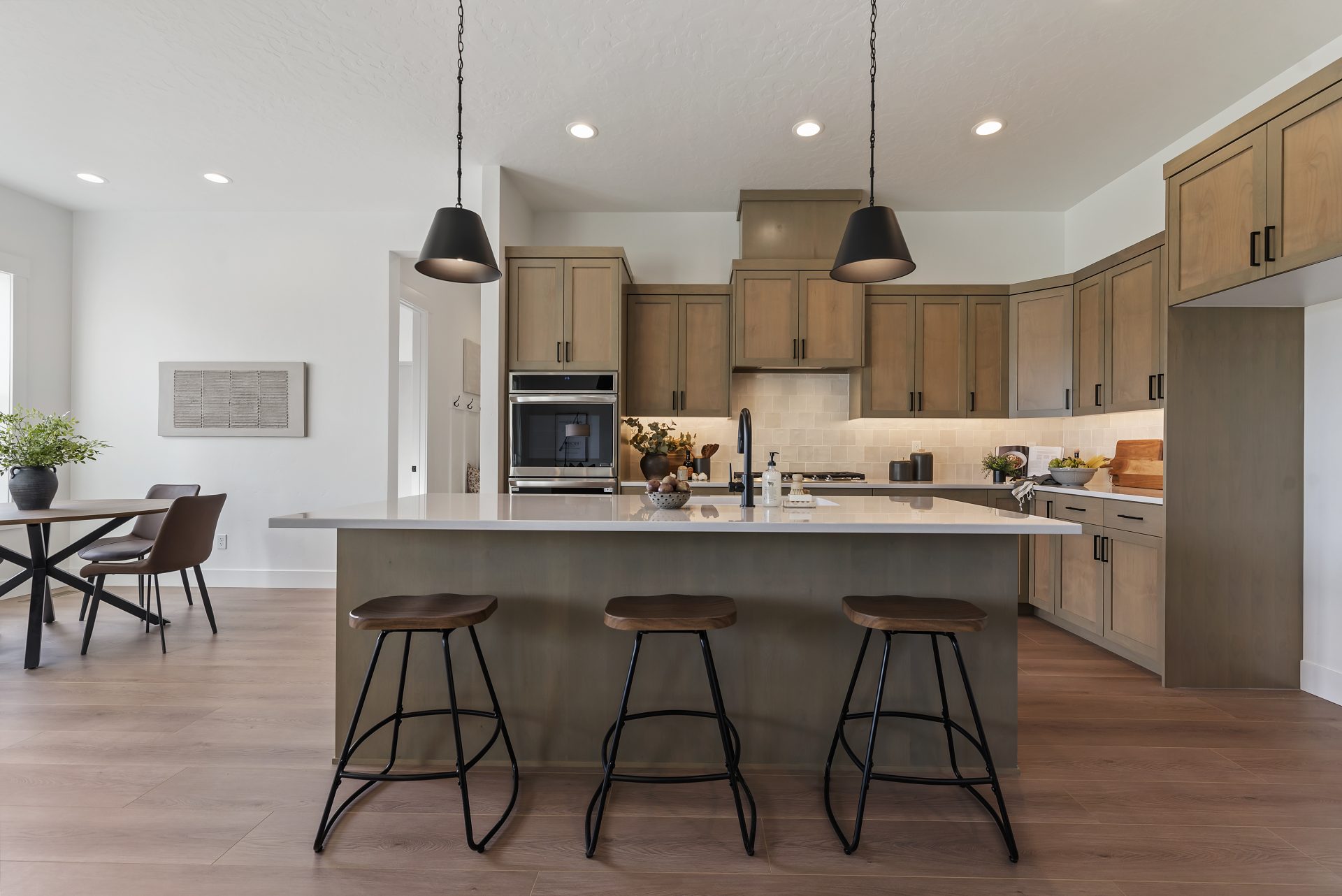 View of a kitchen with an island straight ahead with a brown stained wood base, light color countertop, with a sink  and faucet in the countertop. Behind the island is the view of the cook space with countertop to cabinet tile detail, brown stained wood c