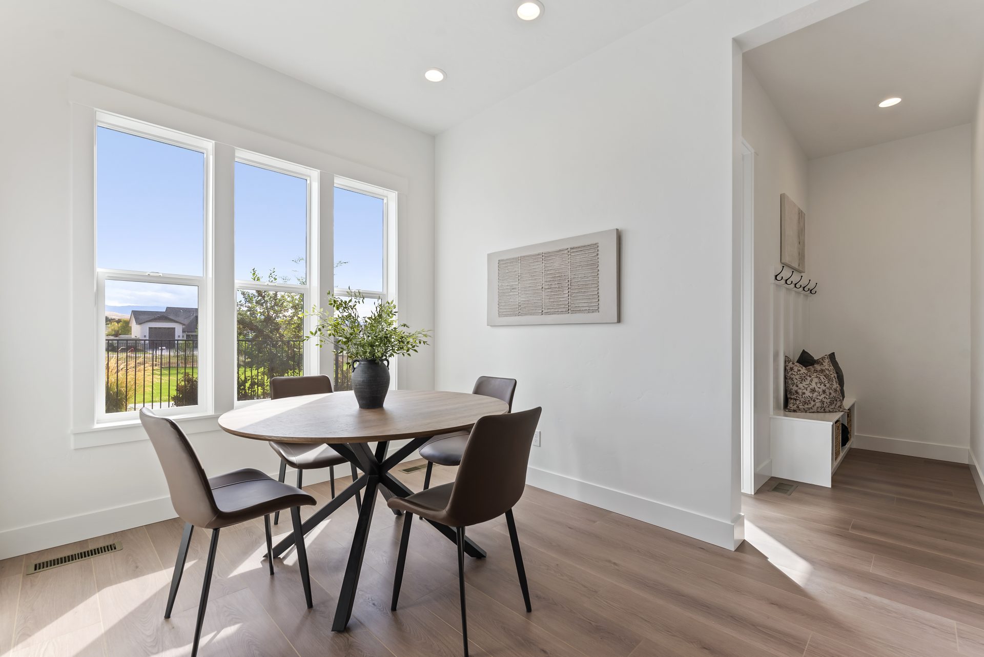 A dining room near three windows, with light colored walls, brown wood texture flooring, a round wooden table with 4 brown chairs and a dark color vase with greenery. To the right a mud room can be seen in the distance.