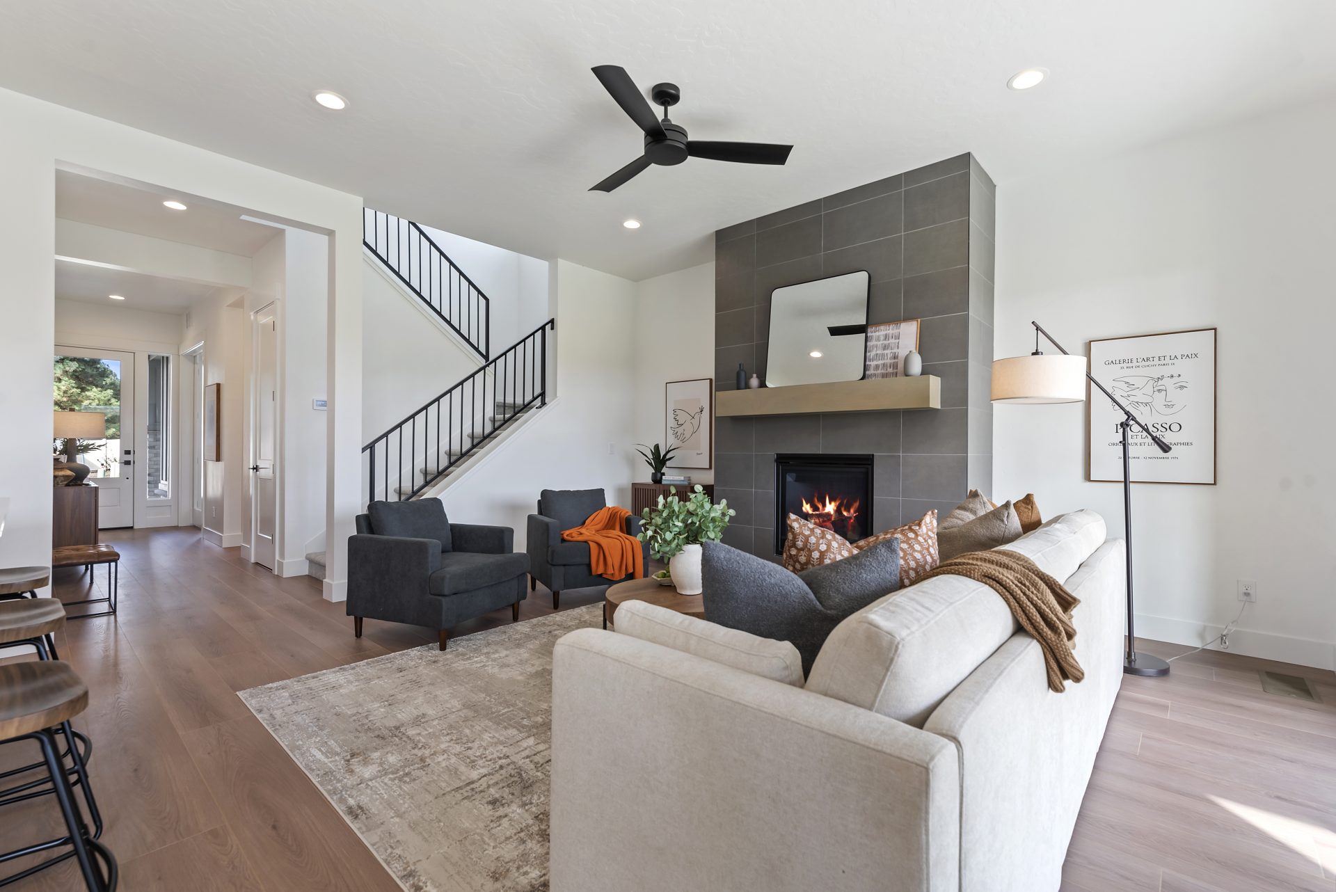 Living room with light-colored couch, neutral pillows, throw blanket, tall floor lamp, two blue cloth chairs, and round wooden table with light vase of greenery. Floor-to-ceiling gray tile surrounds fireplace with wooden mantel. Brown wood-pattern floorin
