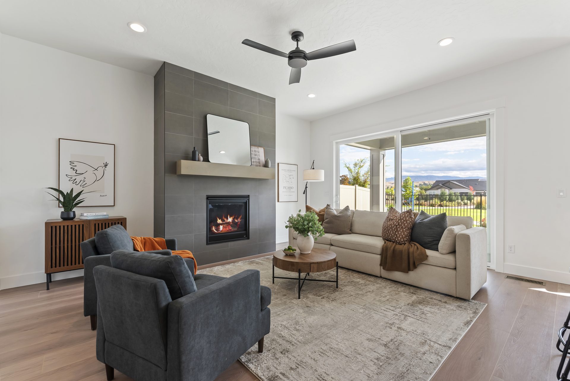 A living room furnished with a light color couch with neutral colored pillows and a throw blanket a tall floor lamp two blue cloth chairs, and a wooden round table with a light vase with greenery.  Floor to ceiling gray tile detail surround a fireplace wi