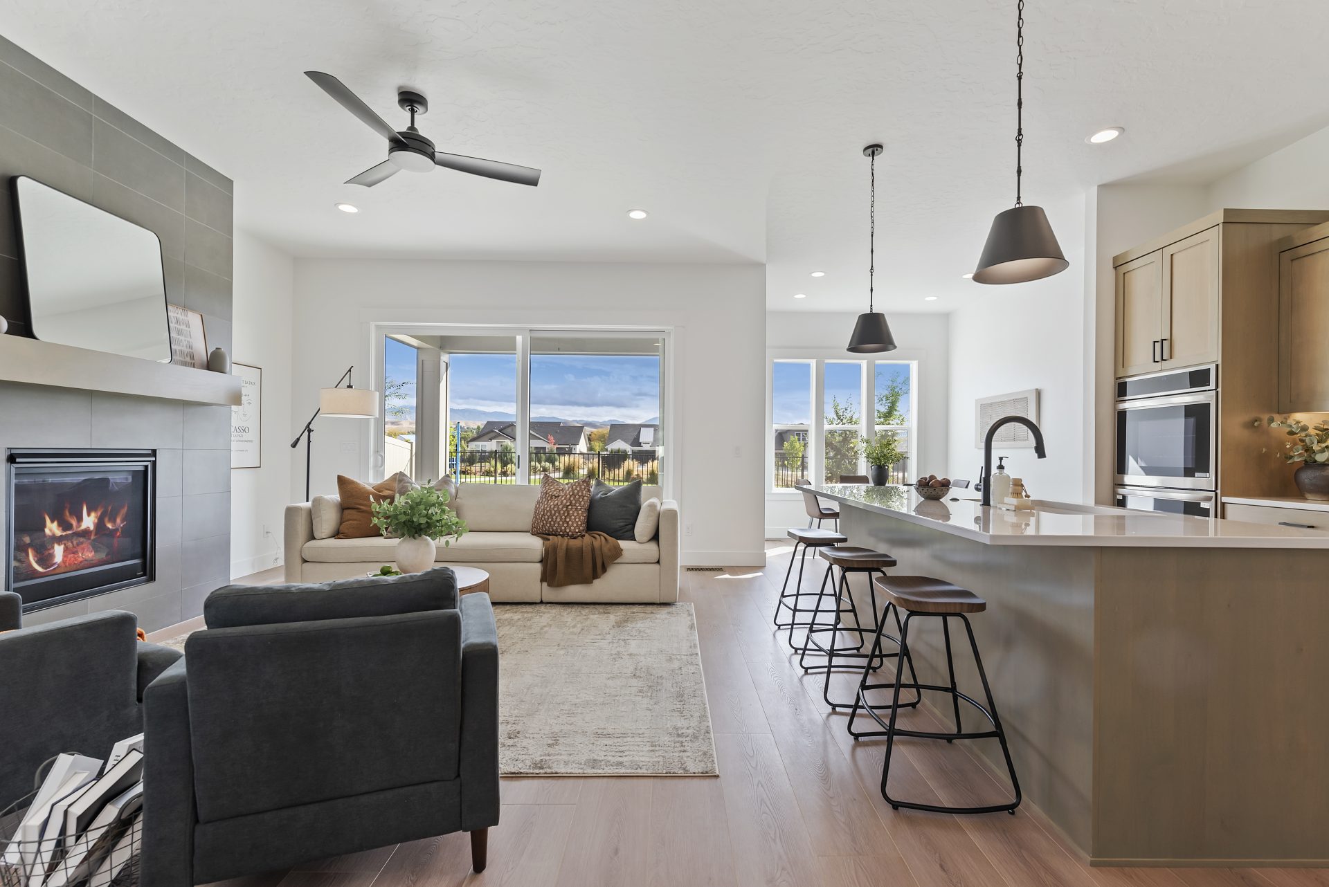 A view of the kitchen and the furnished living room with brown wood- pattern flooring. To the right is the kitchen with brown stained cabinetry, white countertops a large island with a sink and dark faucet. In front of the island are four bar stools. A di