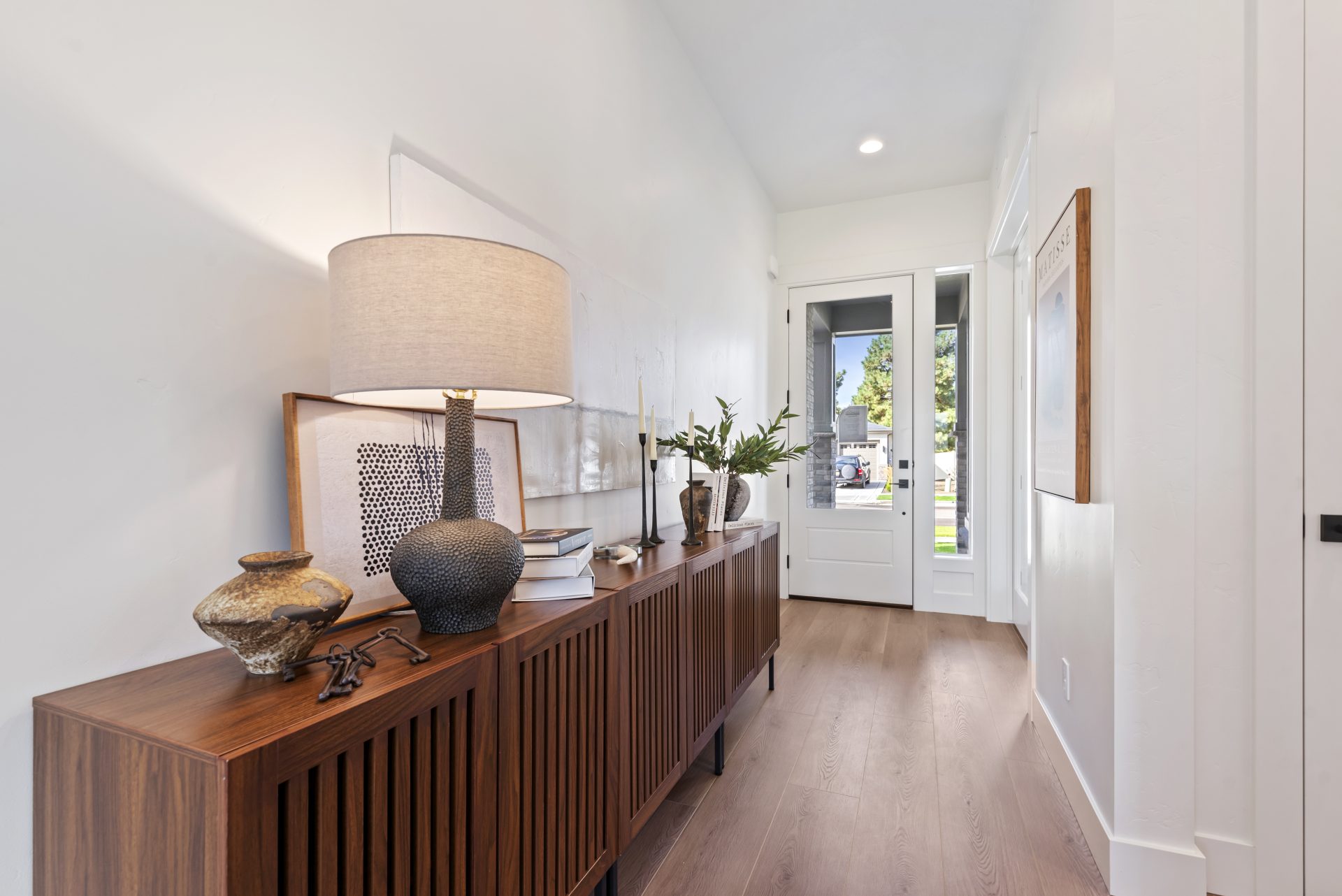 An entry way inside a home with light color walls, brown wood texture flooring, a door with a glass pane, to the left wall an entry wooden table with decorations on top.