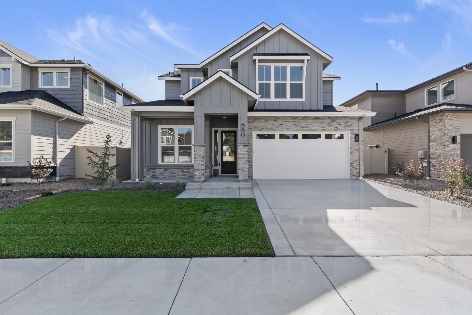 Front view of a modern two-story house with gray paint color, stone accents, and an off-white garage door. The home features multiple windows, a covered entryway, and a neatly landscaped front yard with green grass.