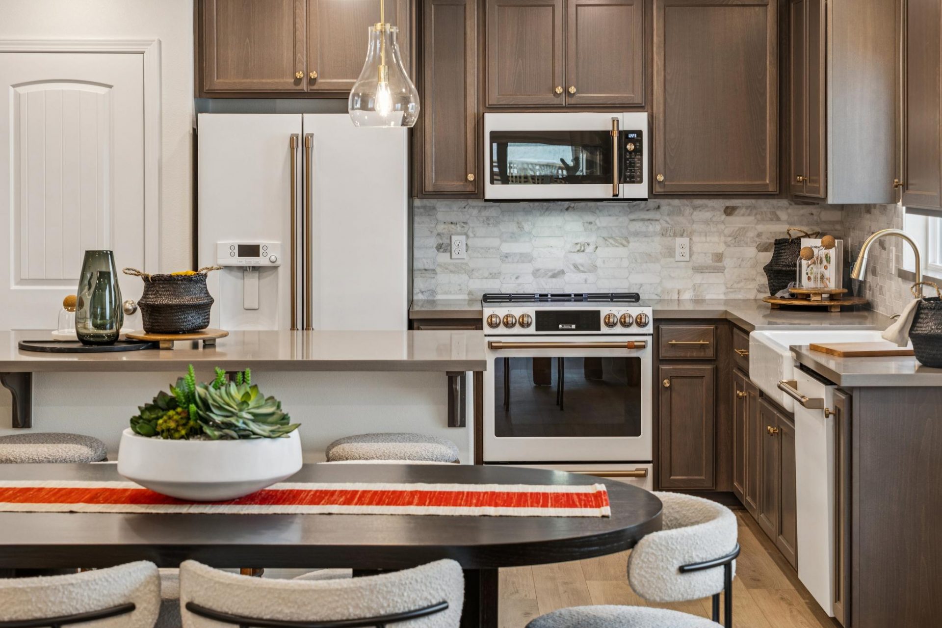 Open concept kitchen with dinning room table, dark beech wood cabinets, dark grey quartz countertops, tiled backsplash, designer white and gold refrigerator and dishwasher, stainless steel microwave and stove, and kitchen island.