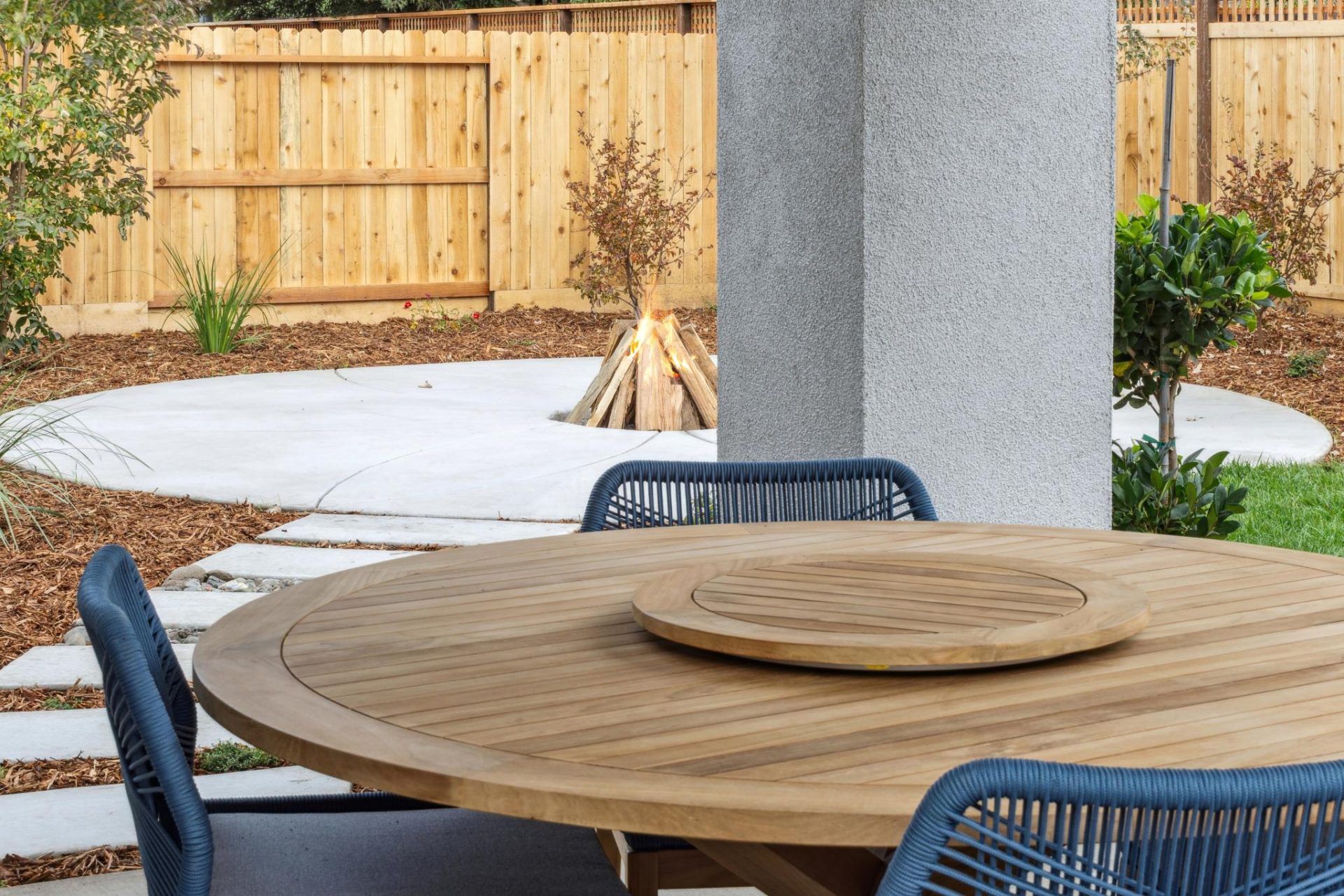 Outdoor oak kitchen table with 3 outdoor chairs in a landscaped backyard with fire pit in the background.