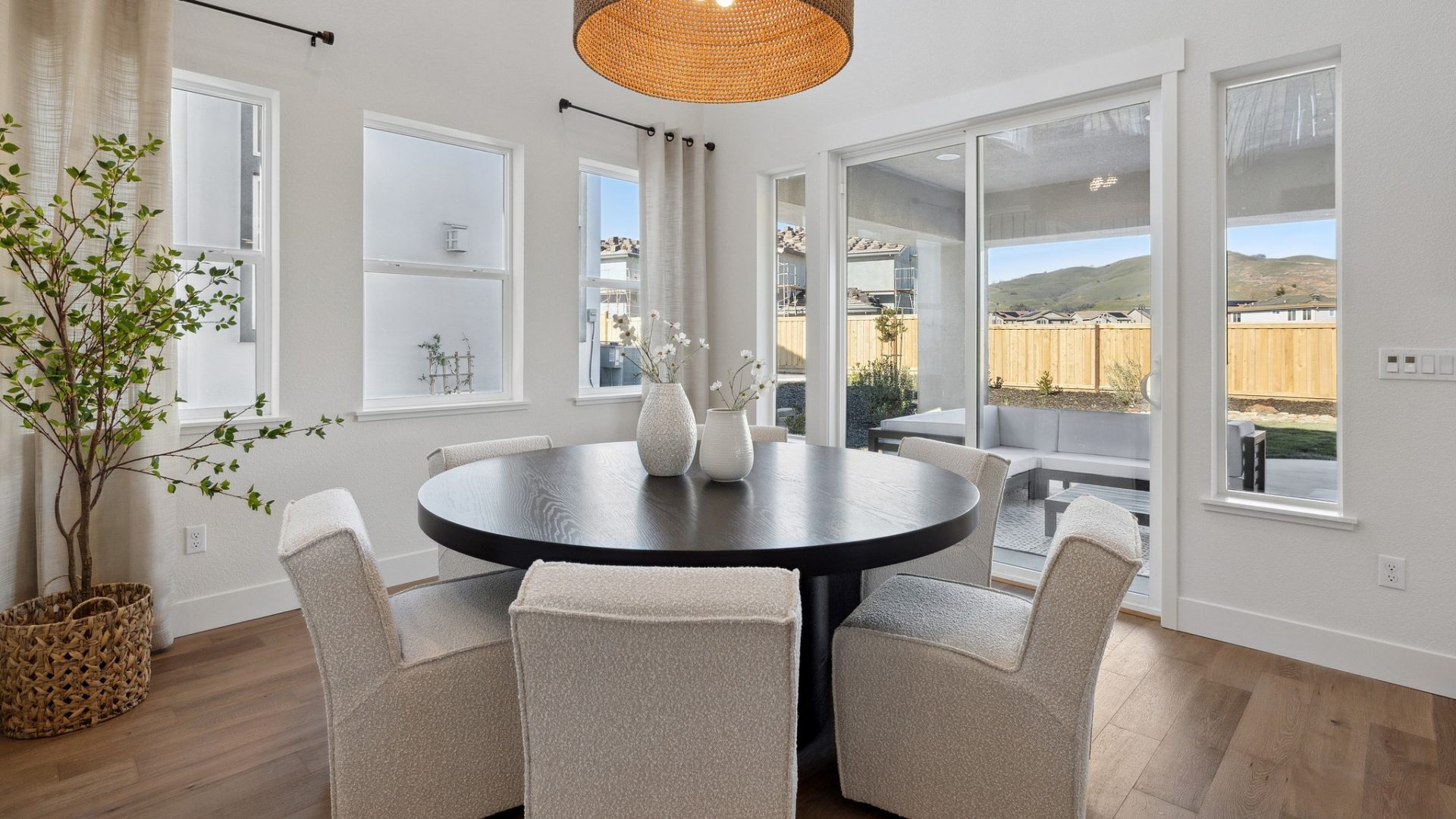 A dining room with a medium round table stained a dark color, with 6 cloth chairs surrounding it and two vases with flower details on top of the table. Behind the table is a glass door slider and multiple windows, and the backyard can be seen.