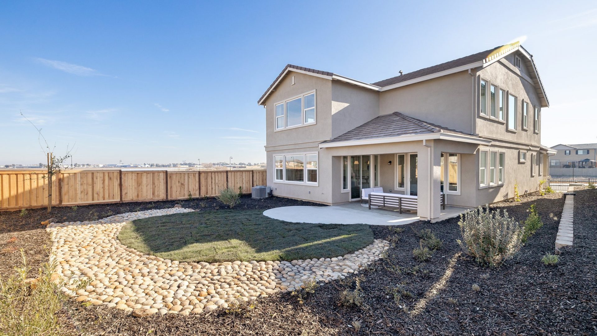 The view of a backyard with river rock, dark colored bark mulch and detailing around a smaller patch of grass. Near the home is a covered patio with outdoor furniture.