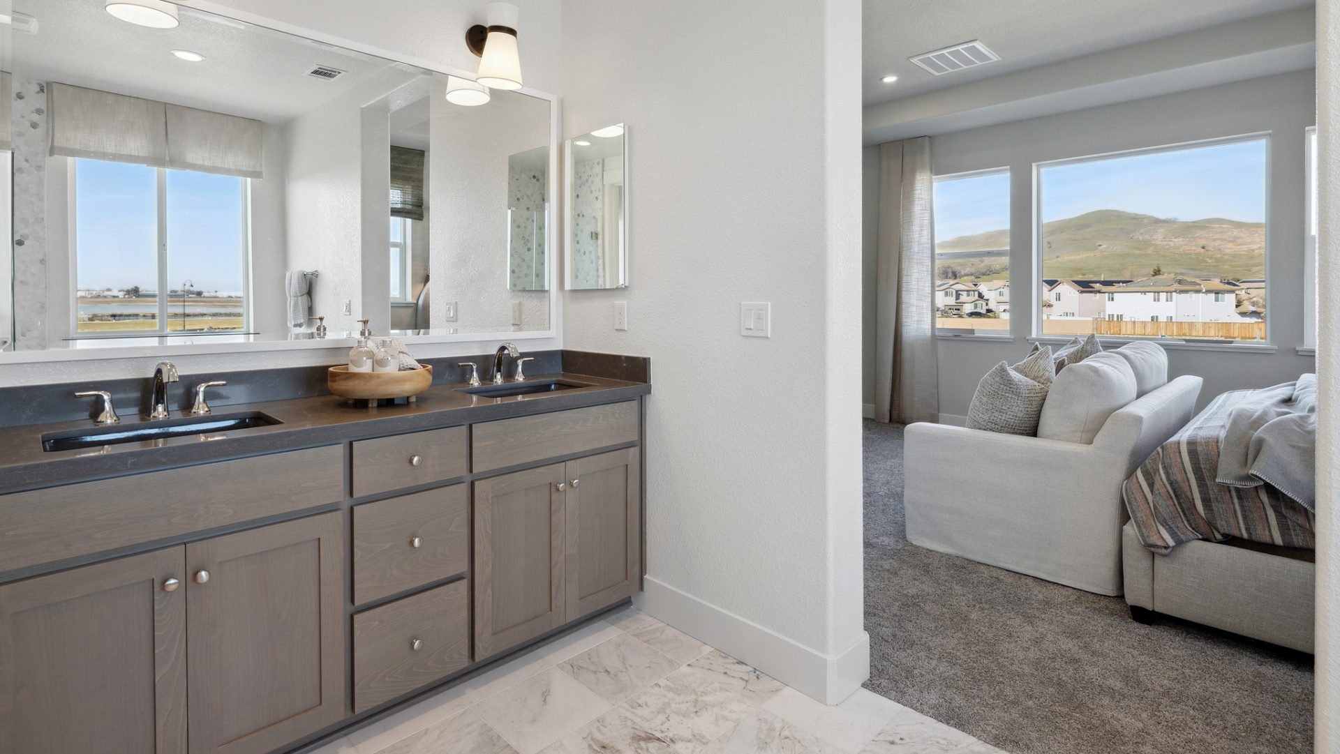 A partial view of a bathroom and a bedroom. In the bathroom is a double sink vanity with darker stained cabinets and a dark colored vanity top with chrome faucets. A mirror spanning the size of the vanity covers the wall and two light fixtures are above.