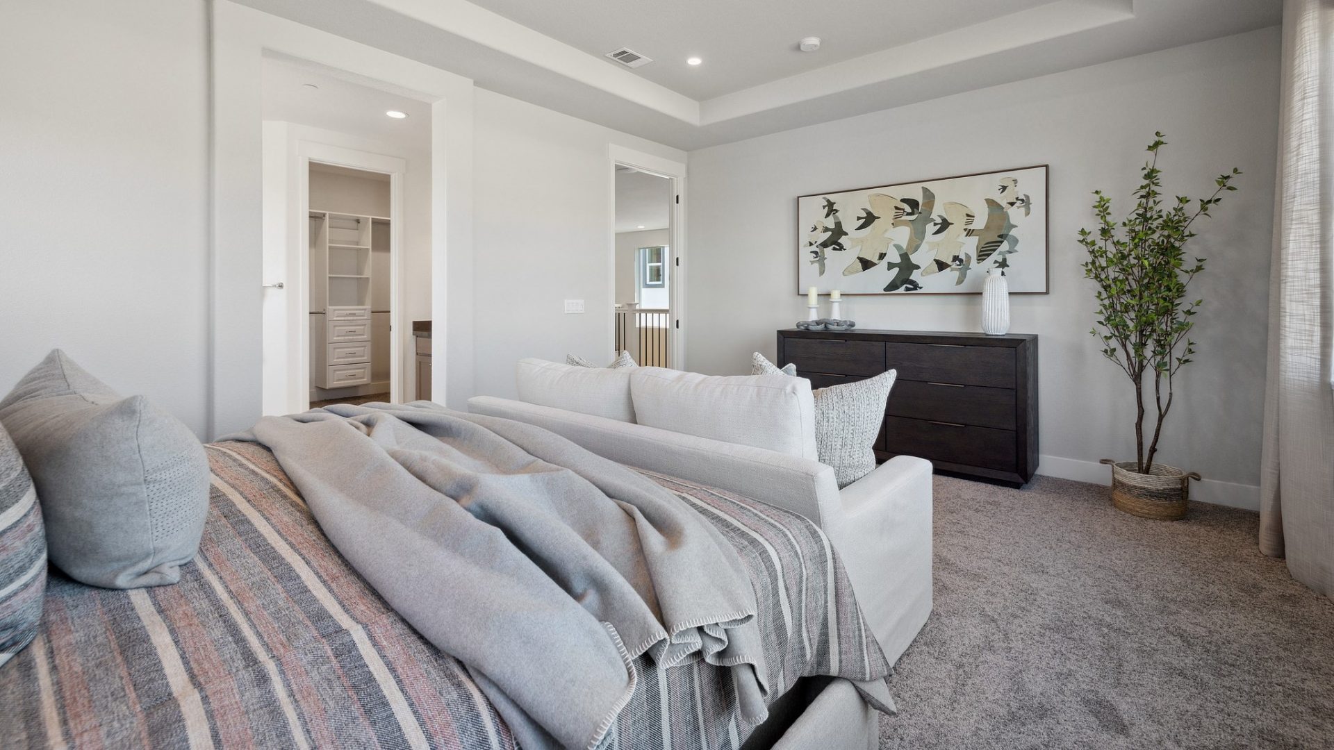 A bedroom with a large bed with grey and reddish multi-color bedding, a dark colored dresser with décor on top and a wall painting located above it with high ceilings and medium gray carpet flooring and other décor items.