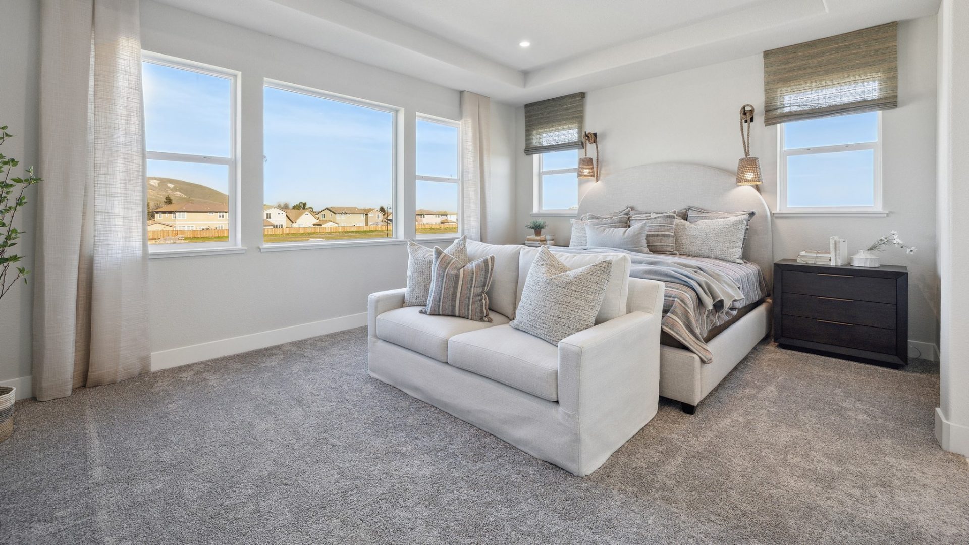 A bedroom with a large bed and a lounge sofa at the end of the bed. A dark stained nightstand on the right. Behind the bed light colored headboard , two windows with window coverings, and to the left are three windows.