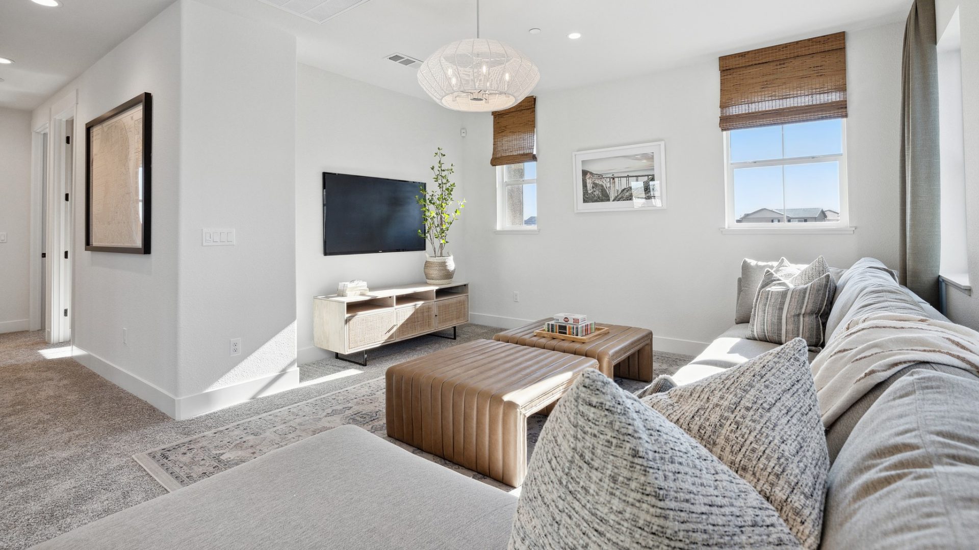 Image of a seating area with light gray color couch with multi-color pillows, and two medium brown coffee tables. In the corner is a short credenza with décor and mounted on the wall above is a tv monitor. To the right are two windows.