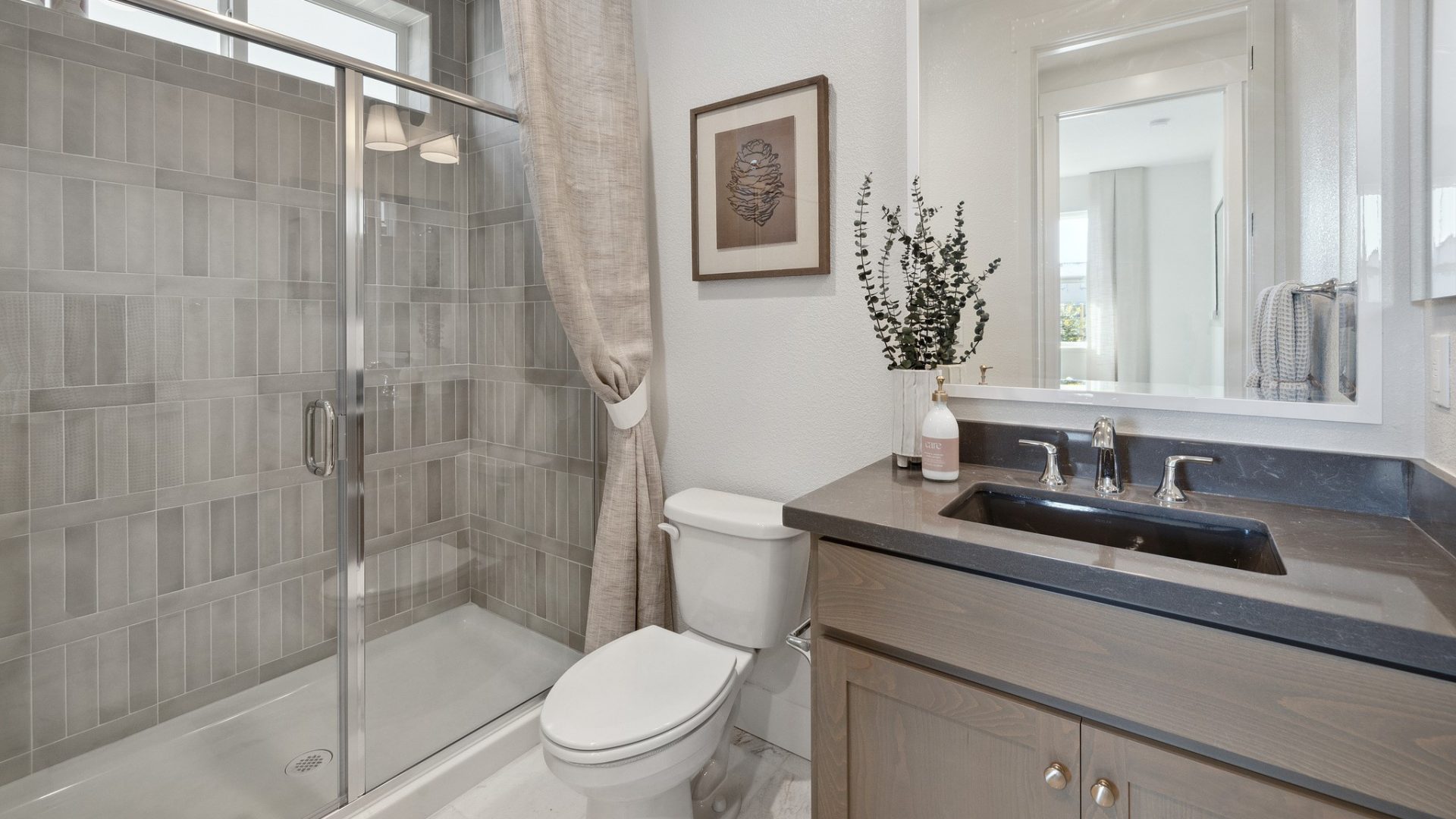 A view of a bathroom with a single sink vanity with a dark stained wood base and dark colored vanity top. A mirror above the vanity and some light décor are located on the vanity top. There is a standing shower with full wall tile and glass surround with