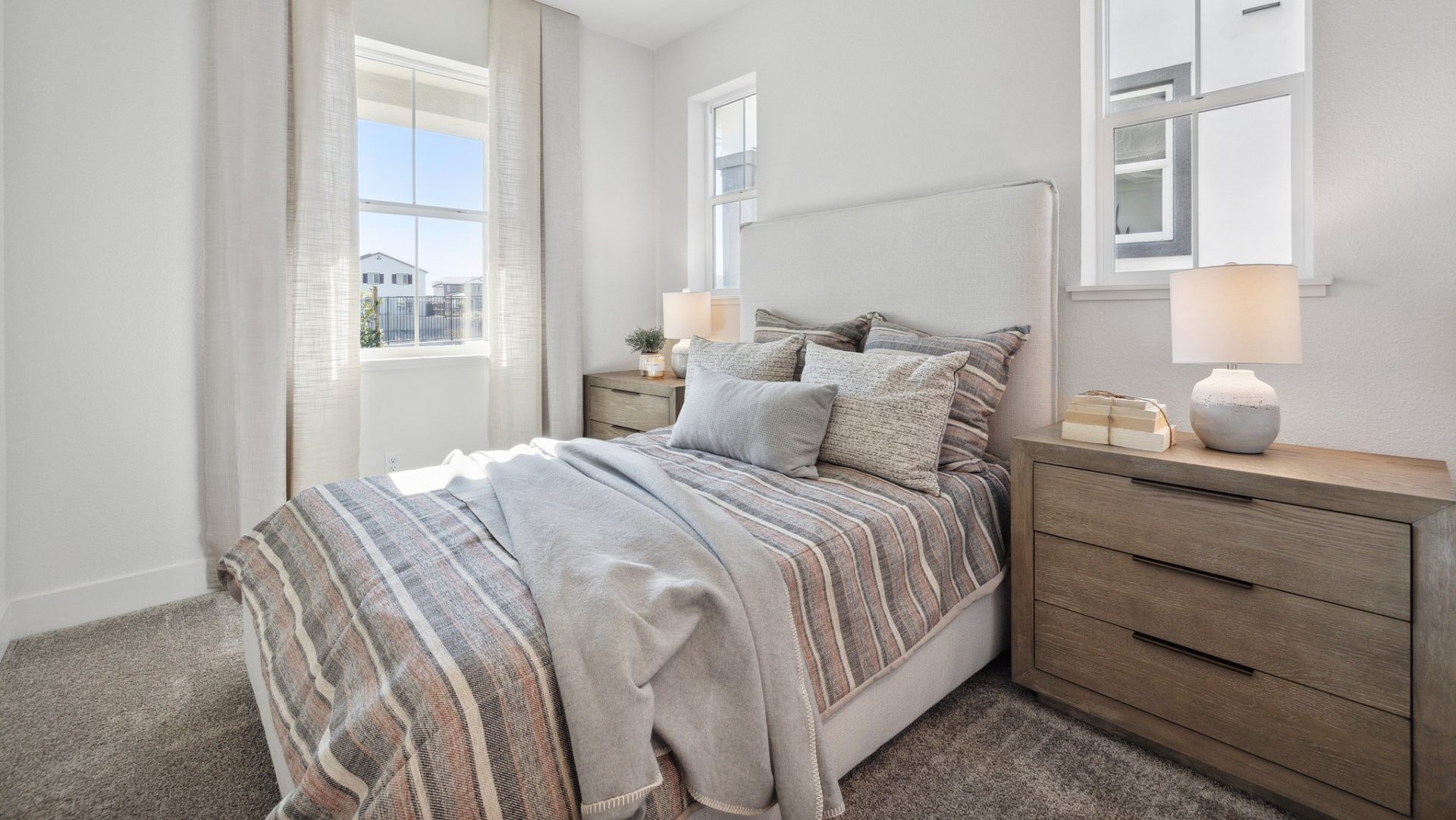 A bedroom with a twin size mattress and headboard with a multi-color comforter and pillows. A medium brown nightstand with a lamp and shade is located on either side of the bed, three windows are located in the room providing ample light.
