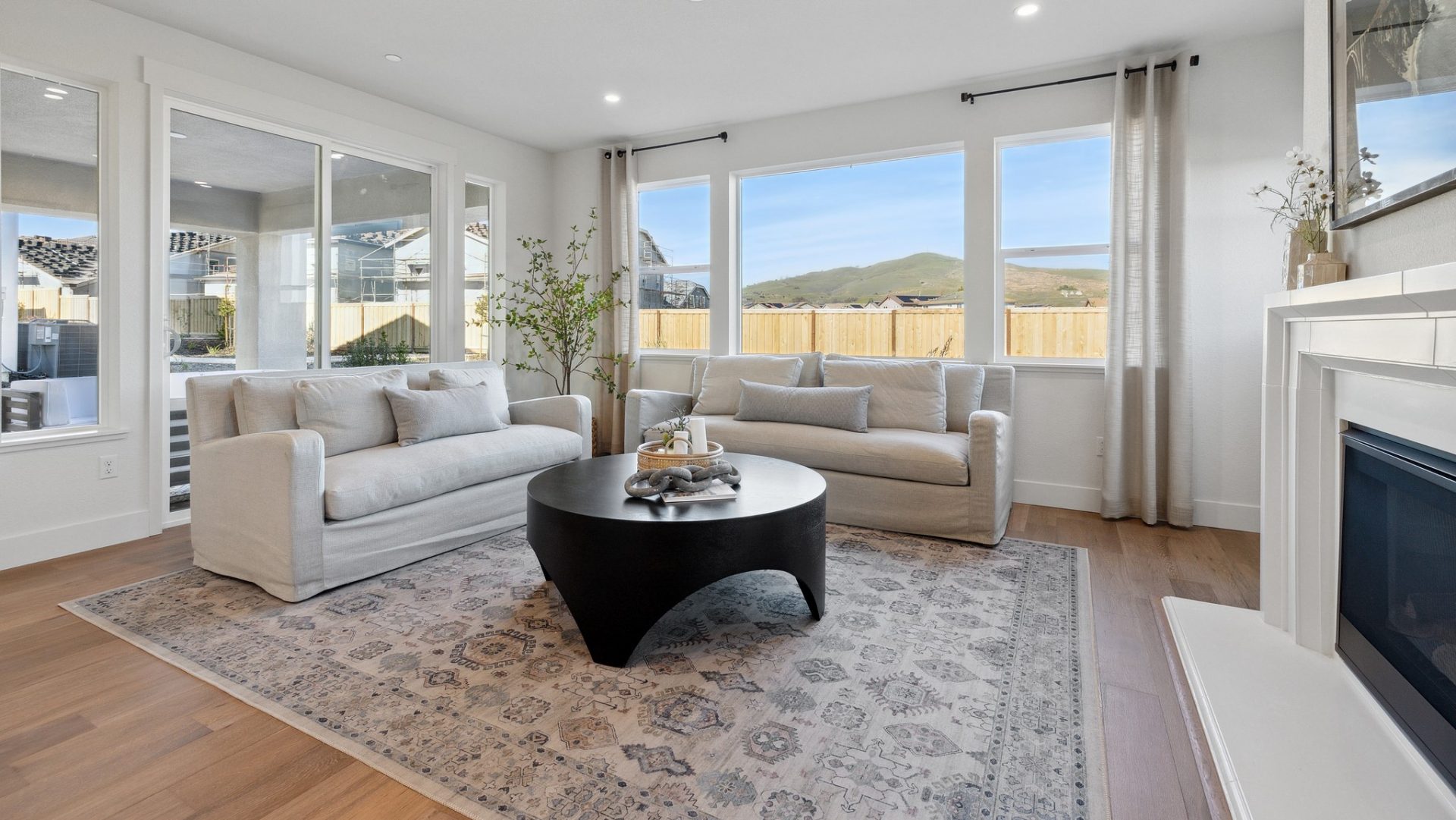 View of a seating area with two light colored couches with light colored pillows, lots of windows providing a bright area, to the right a fireplace with a picture located above and light décor on top of the mantel. In the middle of the room is a large are