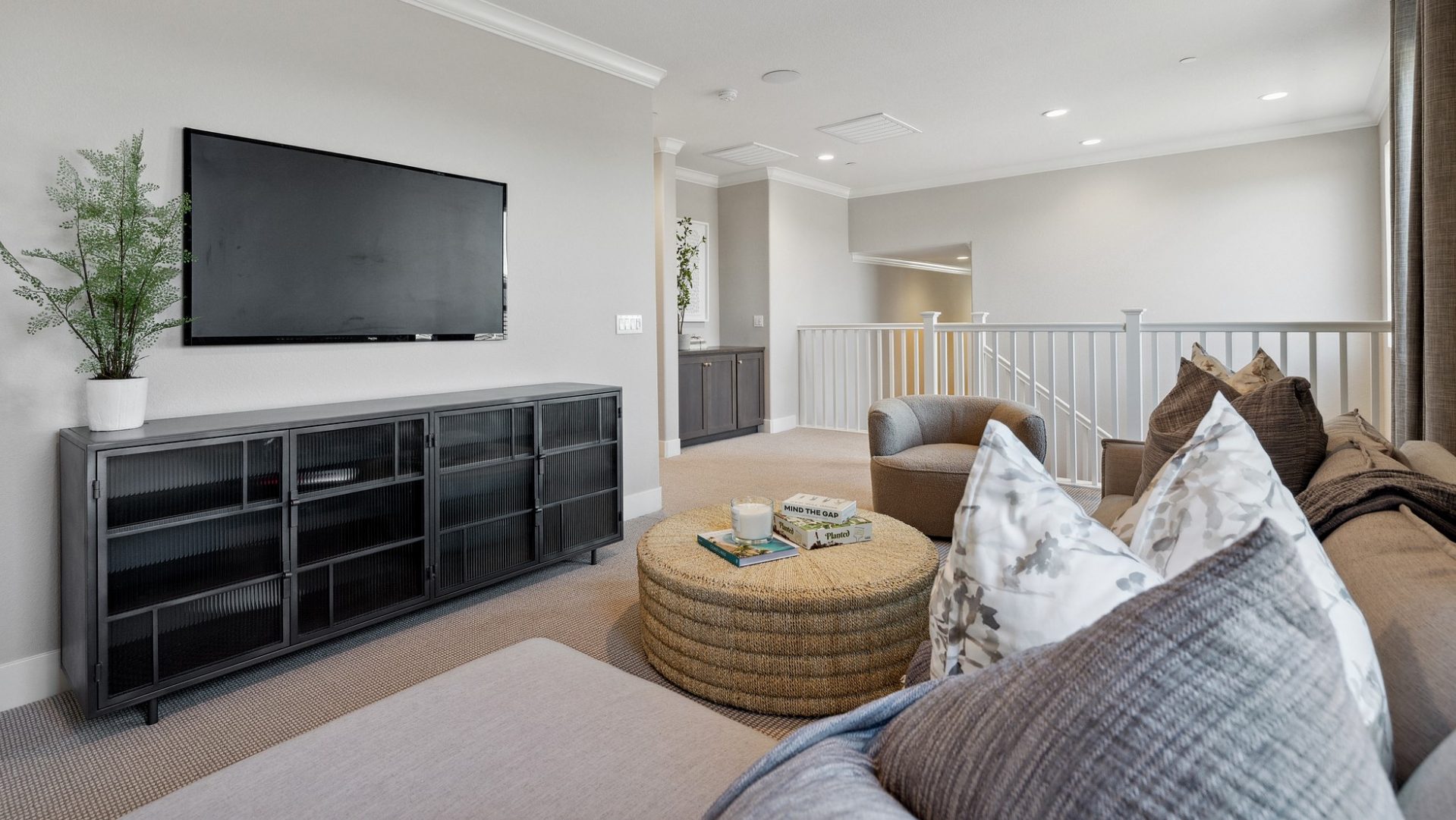 A loft area with a sectional couch with large pillows and a throw blanket, with a round cloth ottoman and a lounge chair. Along the far wall is a dark color credenza with a potted plant on top and a wall mounted tv monitor. In the distance a staircase.