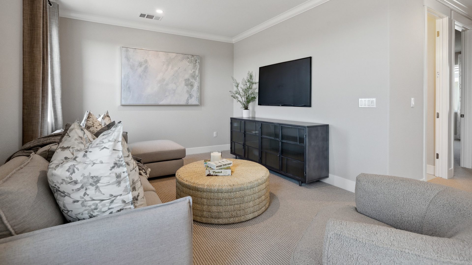 A seating area with a sectional couch with large pillows, and a round tan colored cloth ottoman, a dark colored credenza with a potted plant on top and a wall mounted tv monitor.