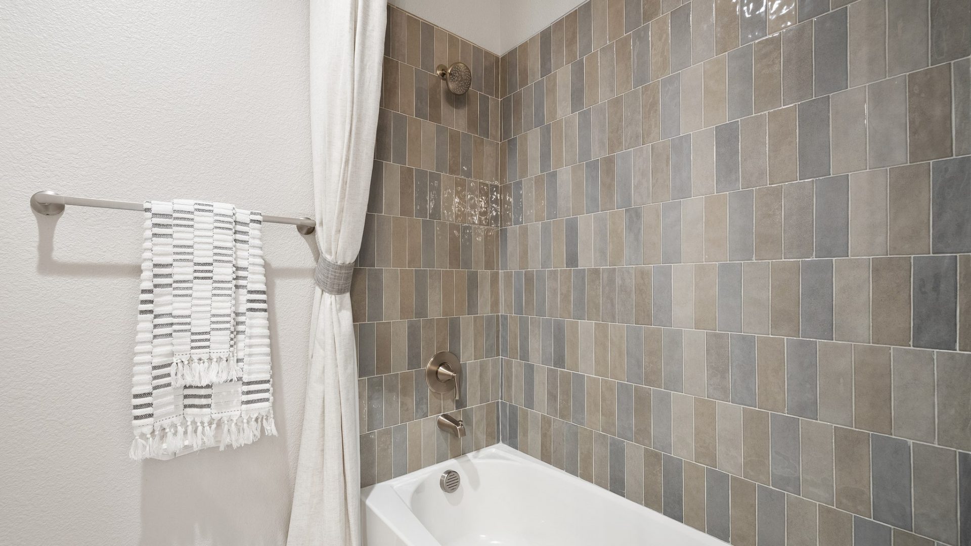 View of a bathtub with full wall of a mix of gray, brown and slate colored tile, with rubbed bronze fixtures and a shower curtain tied back to wall and a towel bar with a towel on it.