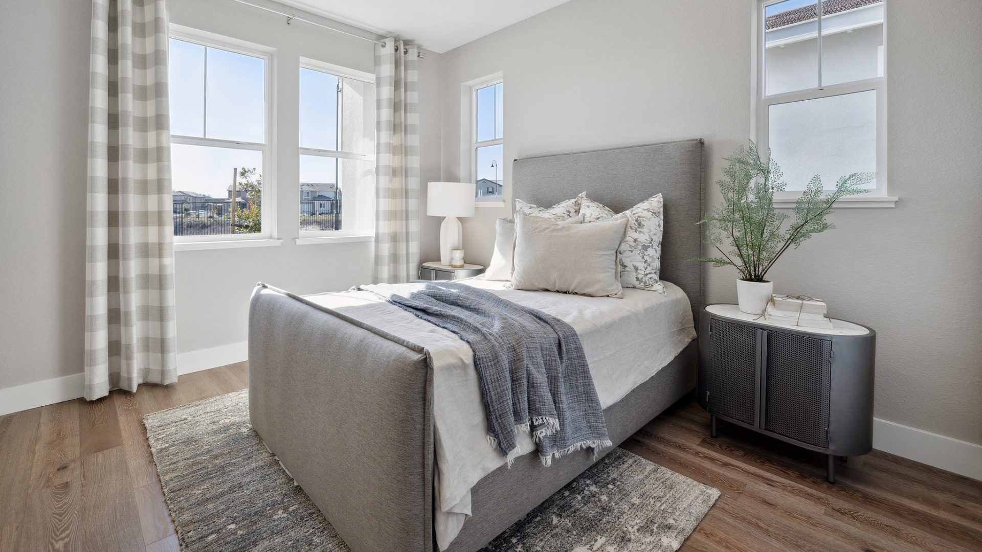 A bedroom furnished with a twin bed in a gray colored cloth head and foot board, area rug and nightstands, with four windows located on adjacent walls, medium brown wood toned flooring.