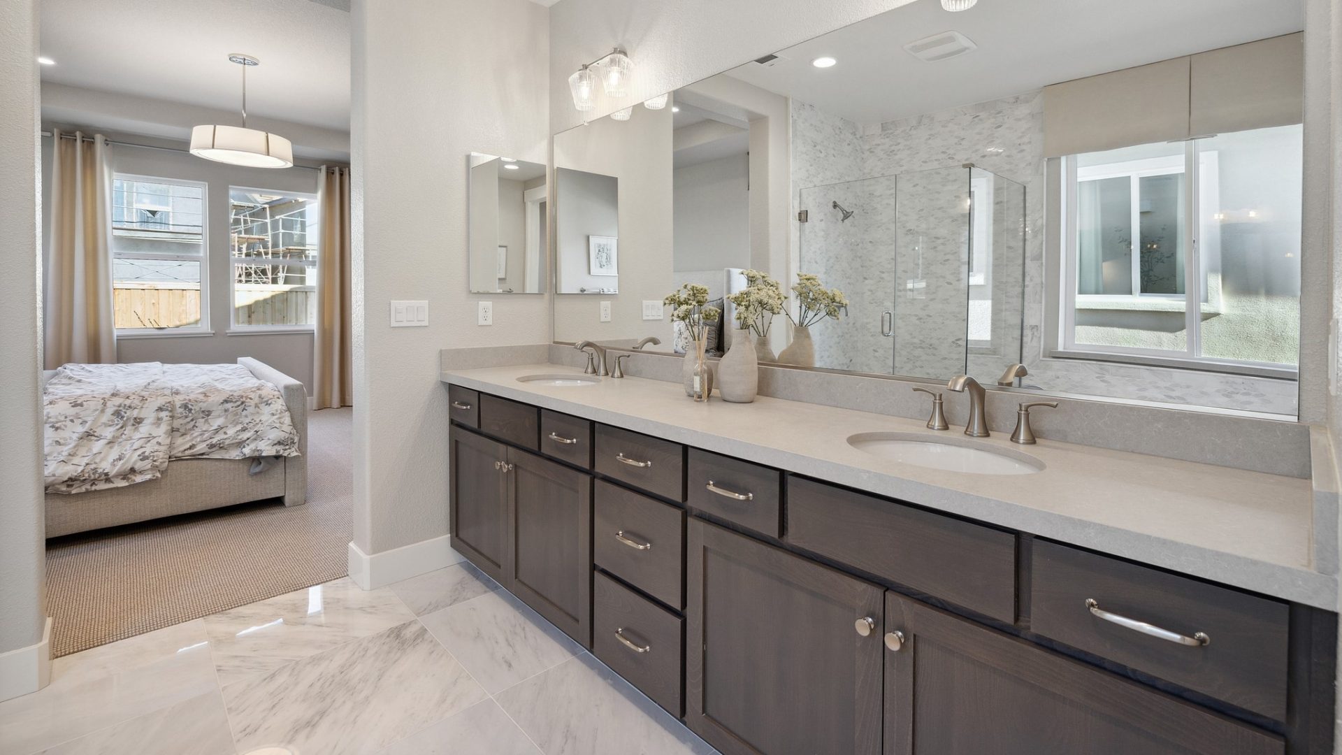 A bathroom with a long dual sink vanity with brushed nickel fixtures and some décor located on top with a long mirror and light fixtures, a bedroom in the distance.