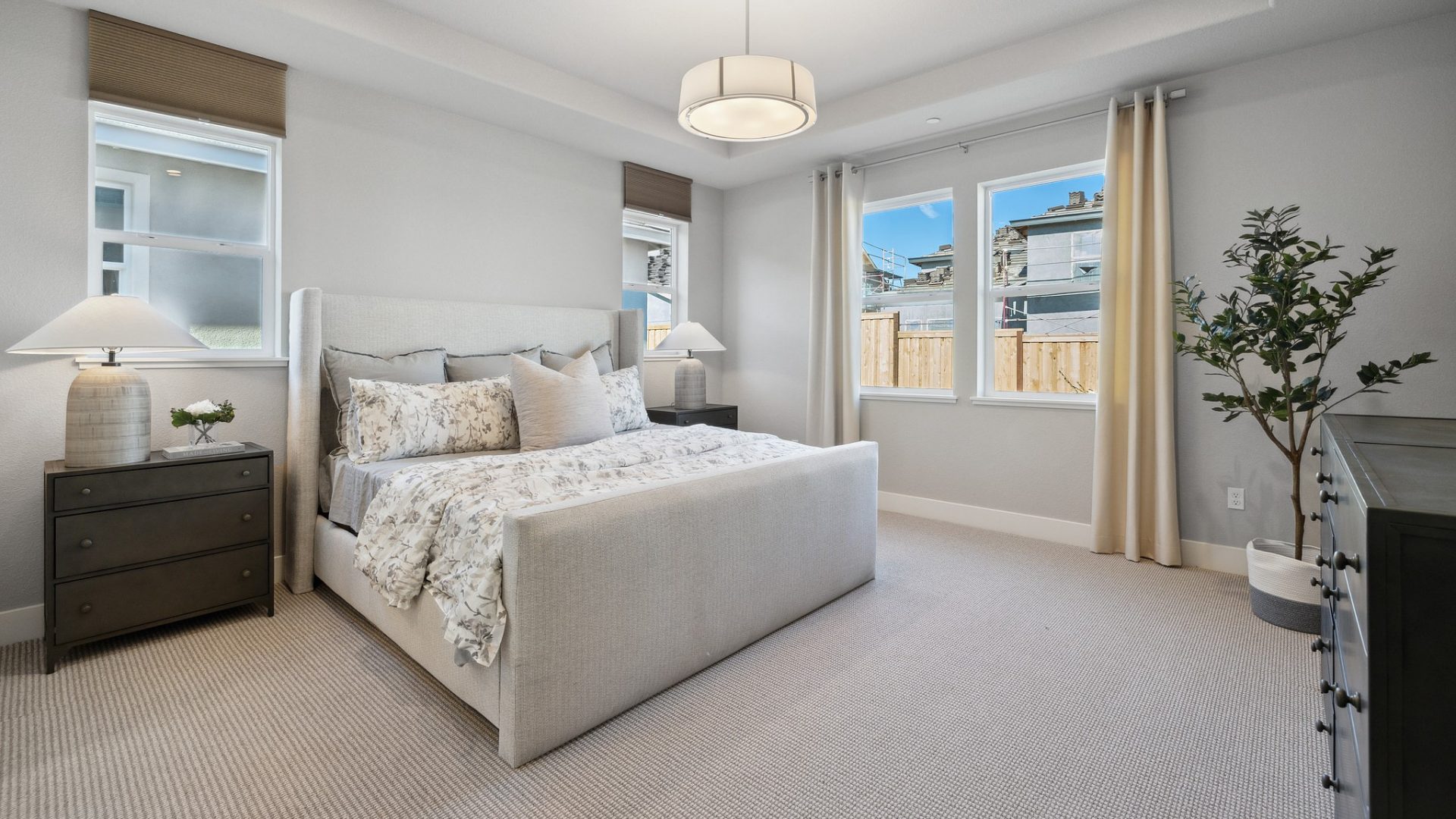 A bedroom with a queen size bed with a light colored cloth foot and headboard with medium brown colored carpet and gray color walls. There are two windows each on two adjacent walls and high ceilings.