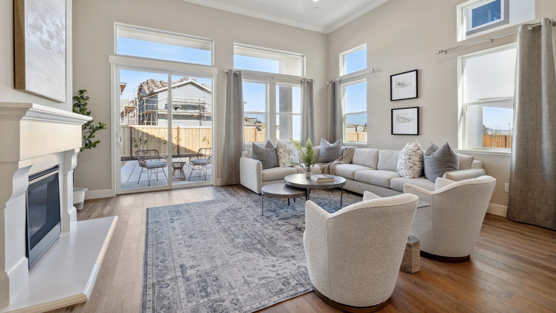 A  living area with a large sectional couch and two cloth seating chairs, high ceilings with tons of windows with hanging curtains. To the left is a fireplace with white details and mantel.