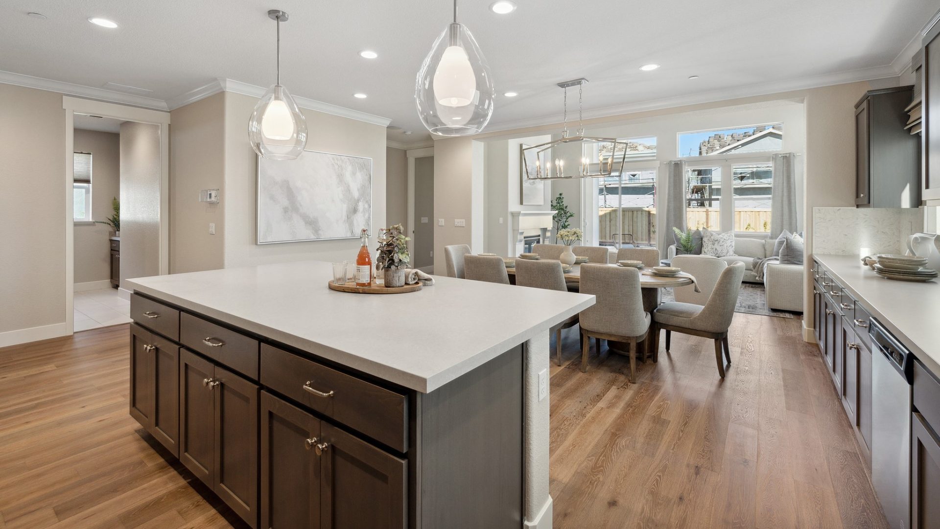 A kitchen island with dark brown stain colored cabinets and drawers with a light colored countertop and a centerpiece located on top. Hanging above the island are two glass pendant light fixtures. Light and medium brown colored wood toned flooring.