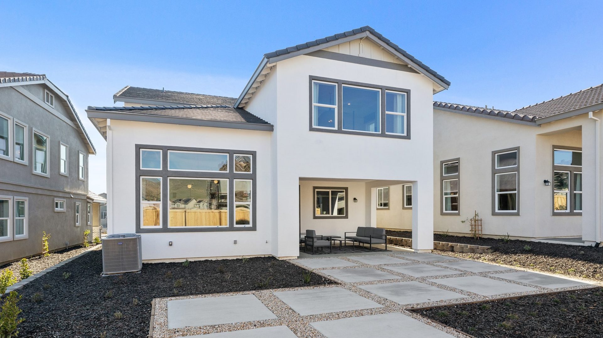 A landscaped and hardscaped backyard with dark colored mulch, a concrete tile pattern pathway with river rock and a off-white stucco house with brown trim and large windows.