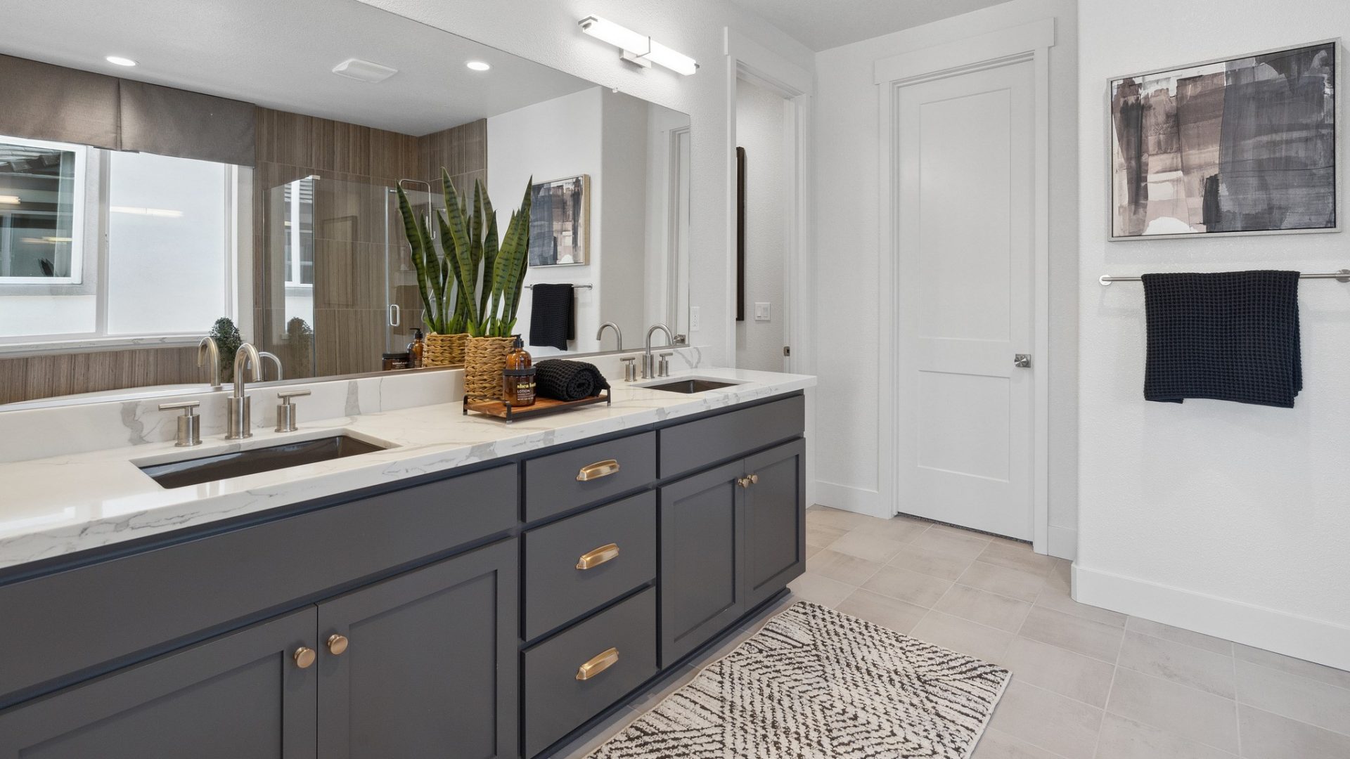 A bathroom with a light color tile floor with a dark gray double sink vanity, with bronze handles on the cabinets and drawers, and light color marble countertop with a plant and other décor items.