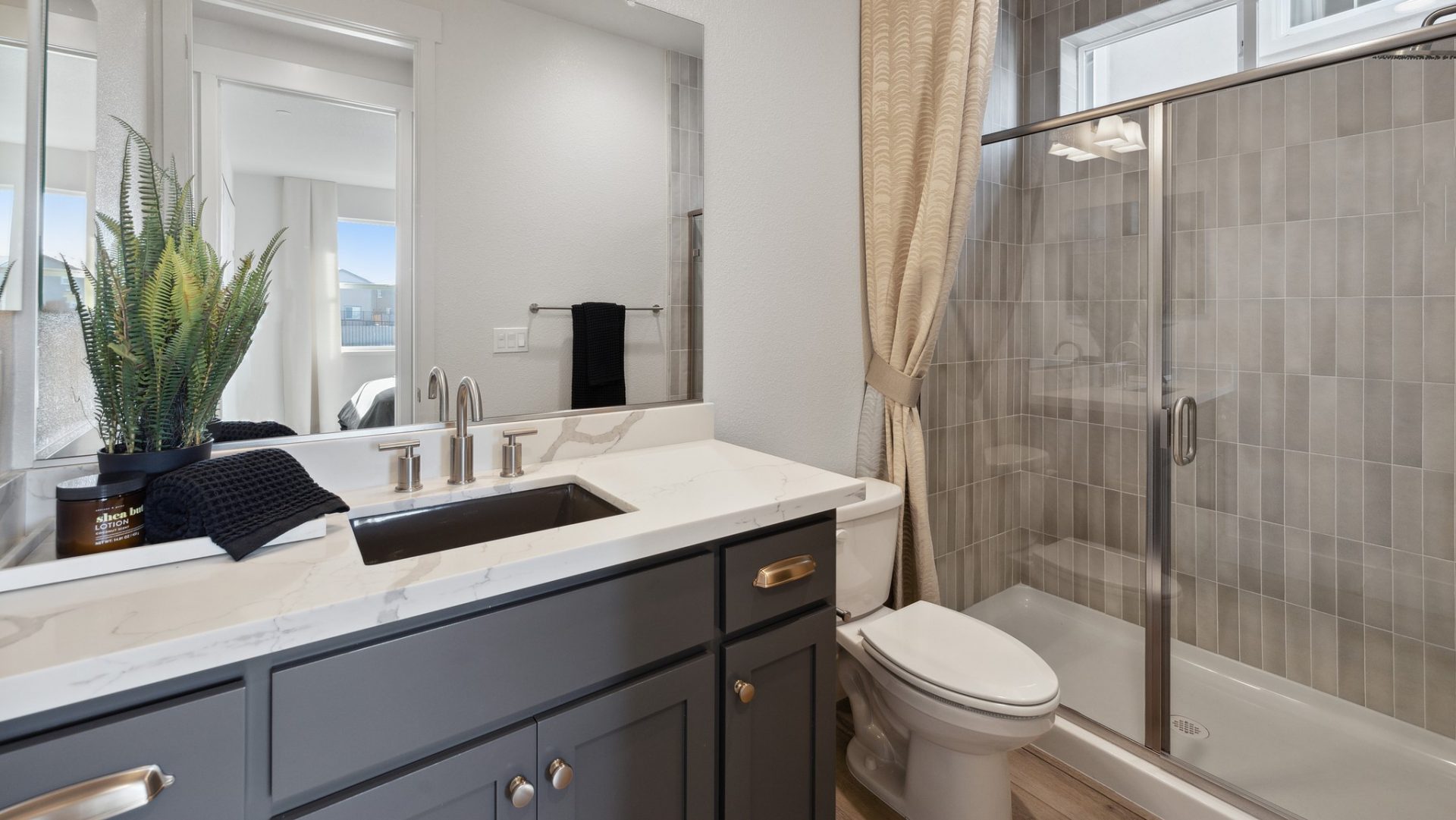 A bathroom with a single sink vanity with a dark colored base and light countertop, a shower with gray tile up to the ceiling and a toilet in between.