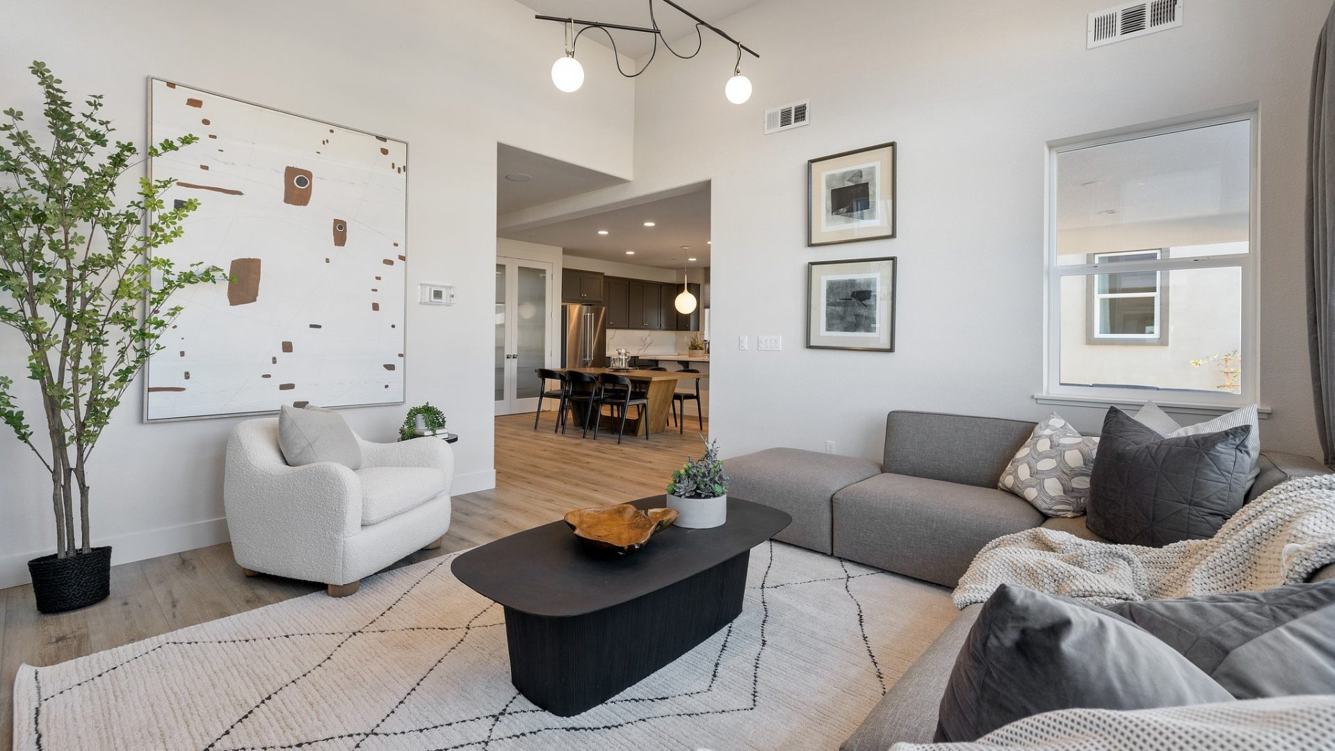 A seating area with gray sectional couch, dark color coffee table, light color lounge chair, high ceilings with a modern light fixture and various décor.