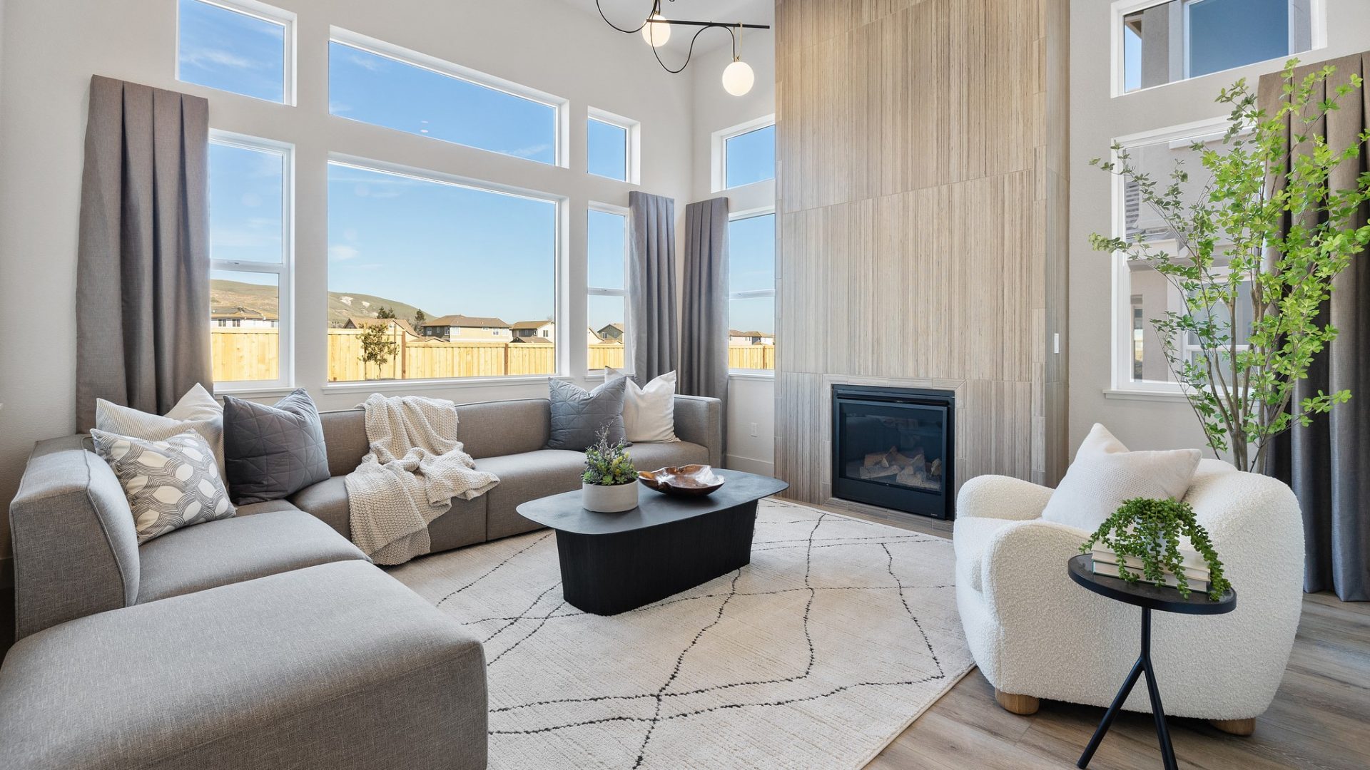 A living area furnished with a sectional couch and lounge chair, a large area rug, with an electric fireplace with tile detail to the ceiling, large windows and high ceilings.