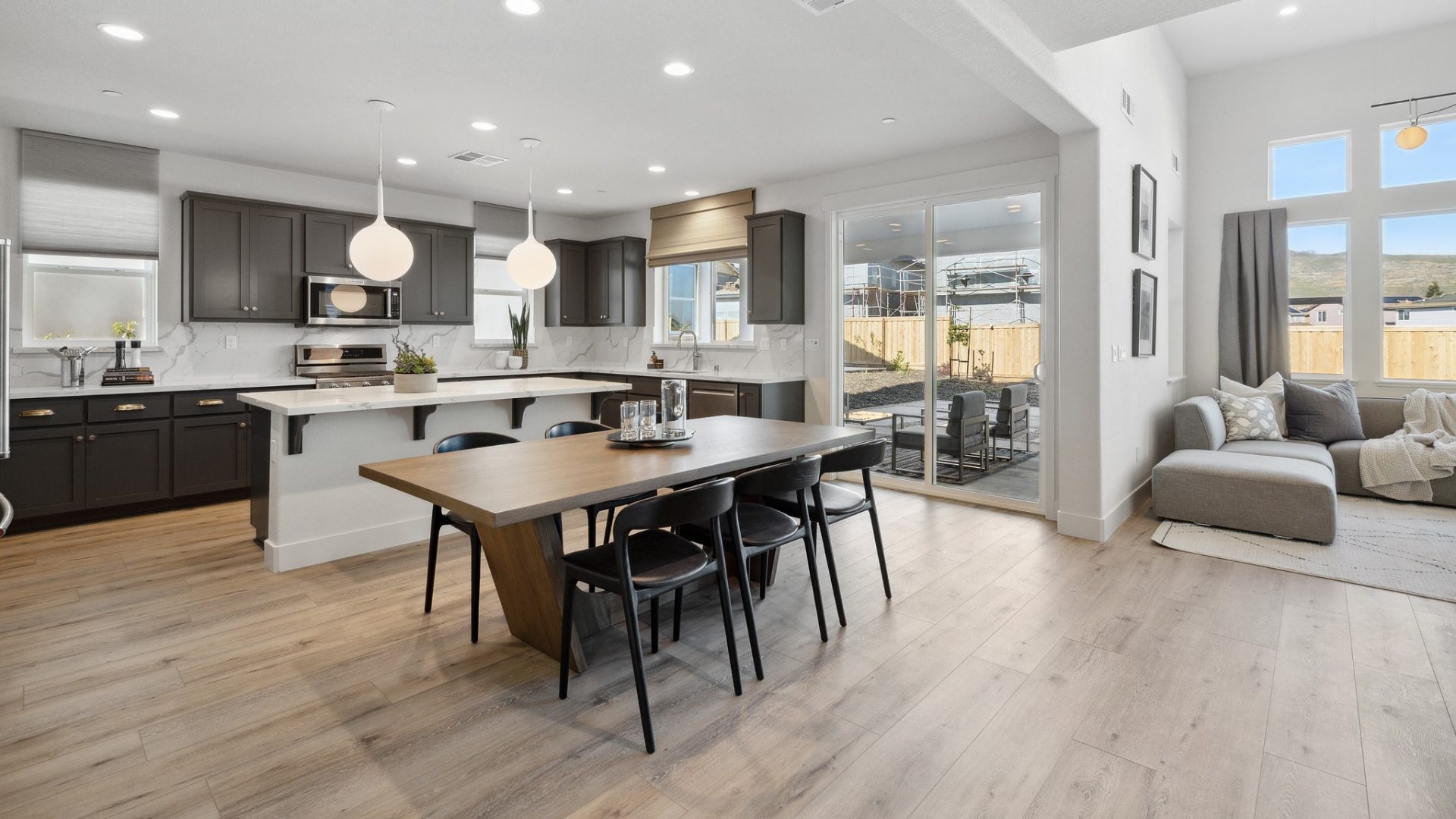 A kitchen and dining area with medium brown wood flooring with windows lining the walls, a sliding glass door and a long dining table with six chairs, kitchen in the background with pendant lighting over a kitchen island.
