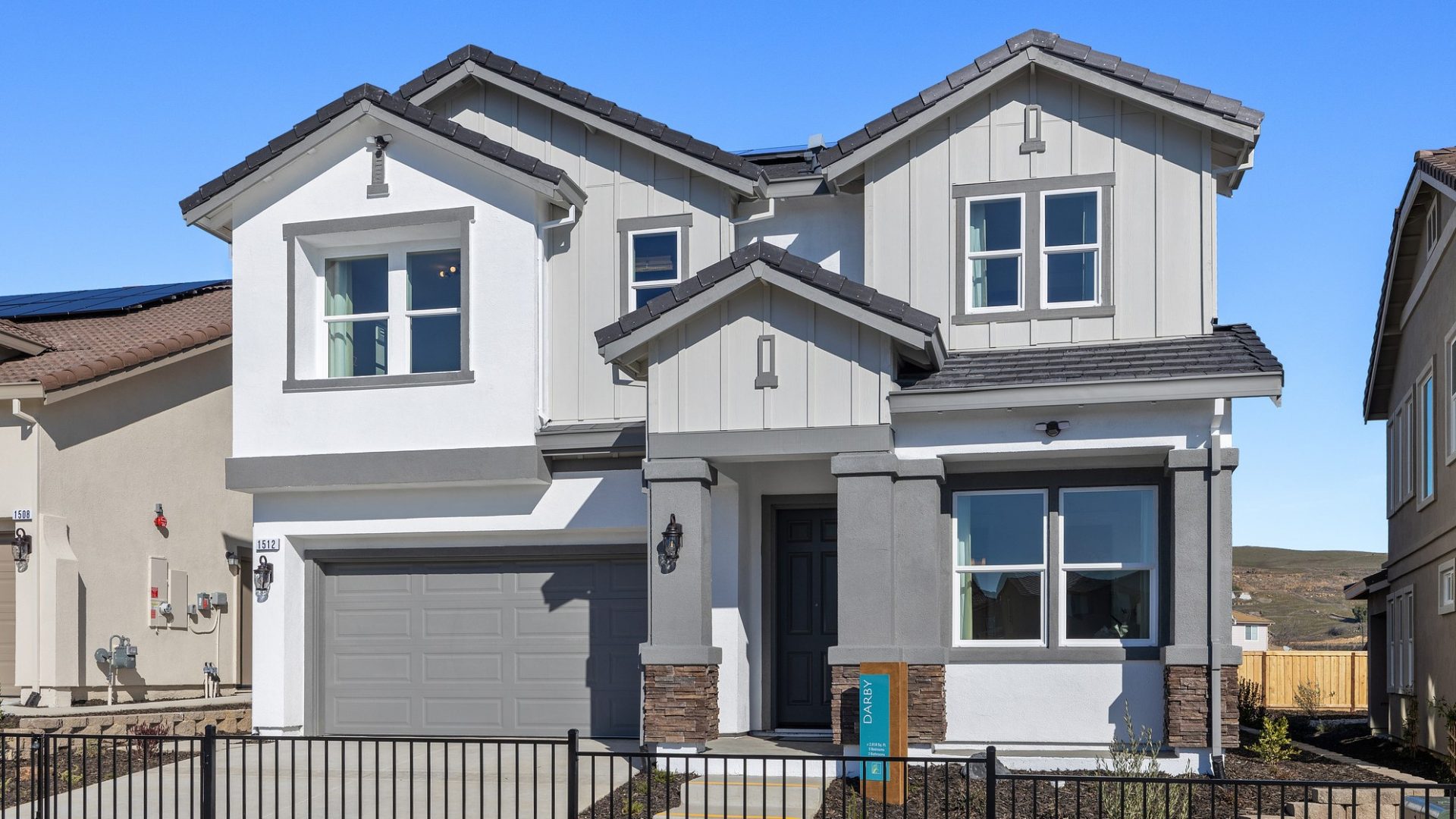 A front exterior of a home with stucco and wood paneling with a three color paint scheme from off- white to medium and dark gray with mulch landscaping with small plants. A tall covered entry way with pillars that have brown stone accents at the base.