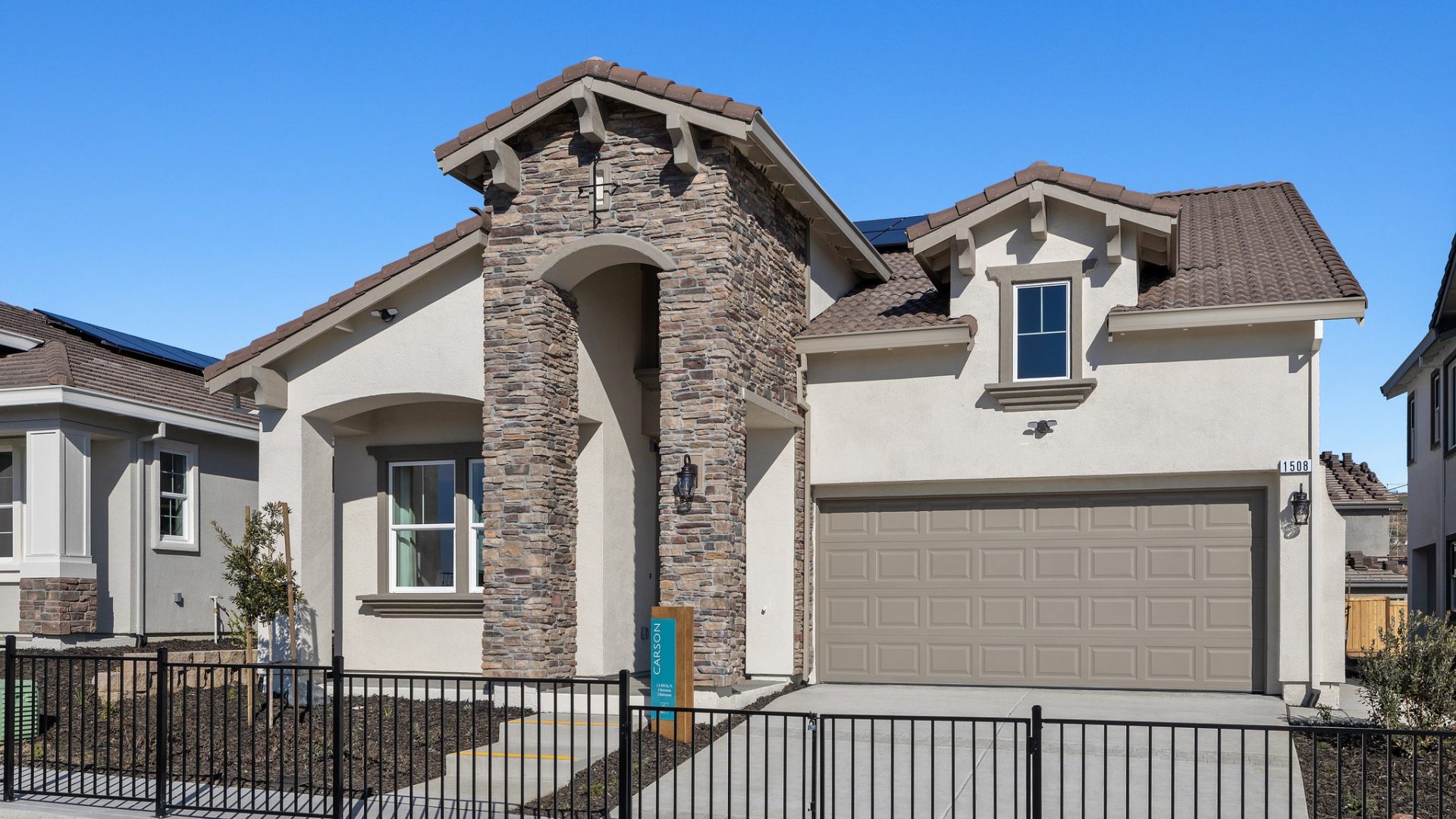 The exterior of a home with light tan stucco with brown color accents, a large entry way with a ground to roof stone detail of mixed colored stone with dark colored mulch and small trees and shrubs landscaping.