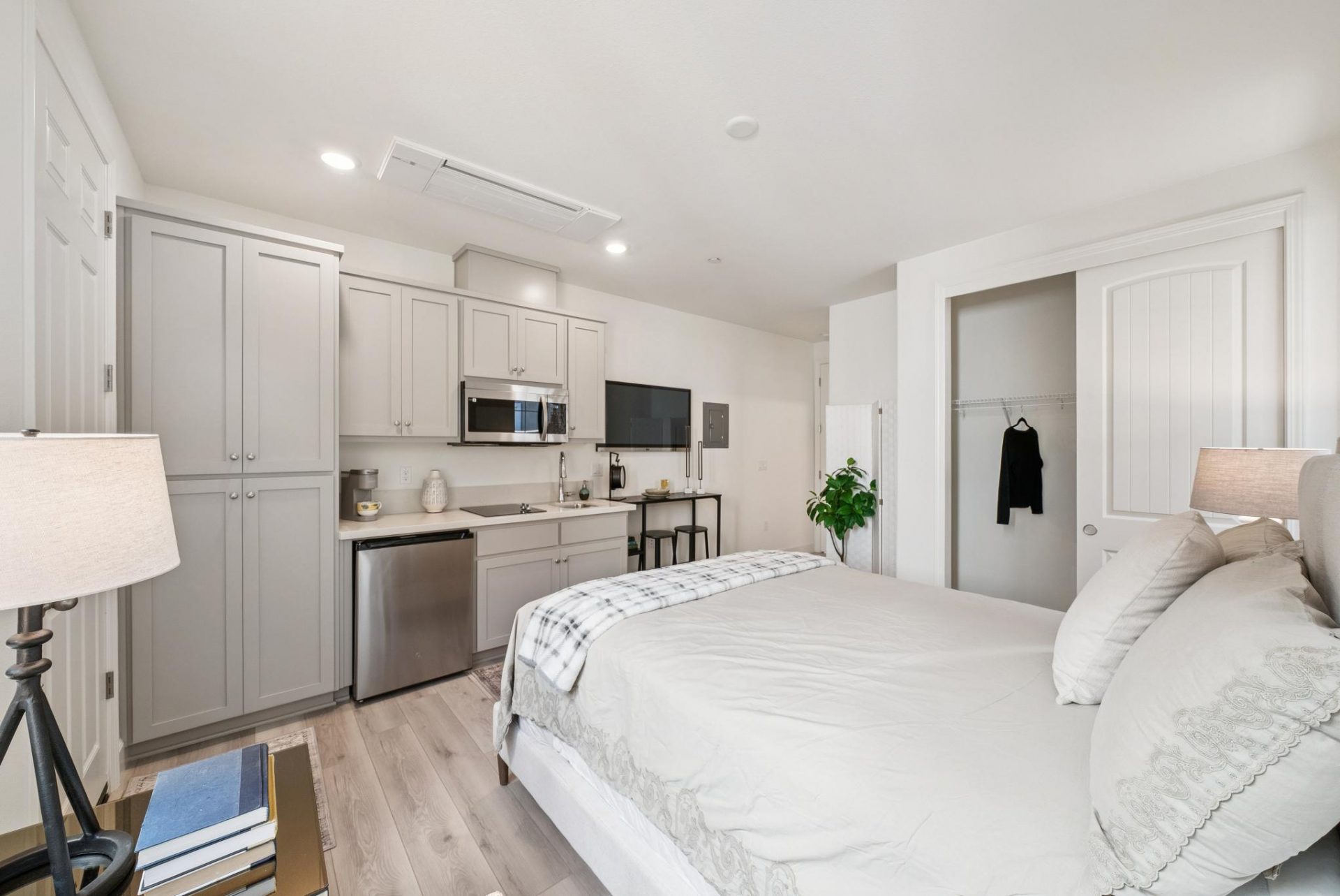 Room furnished with a bed, a kitchenette with a dishwasher, microwave and stovetop ,cabinetry and countertop space. Light gray wood colored flooring and a sliding door closet.