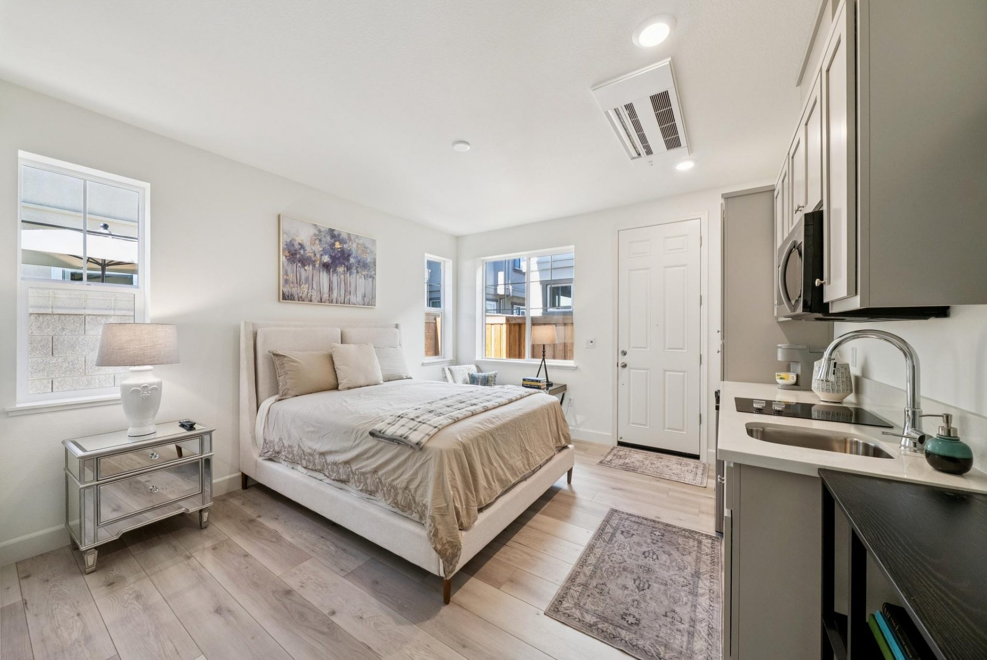 Room furnished with a bed, a kitchenette off to the right, around the bed are three windows, and a nightstand with a lamp on it.