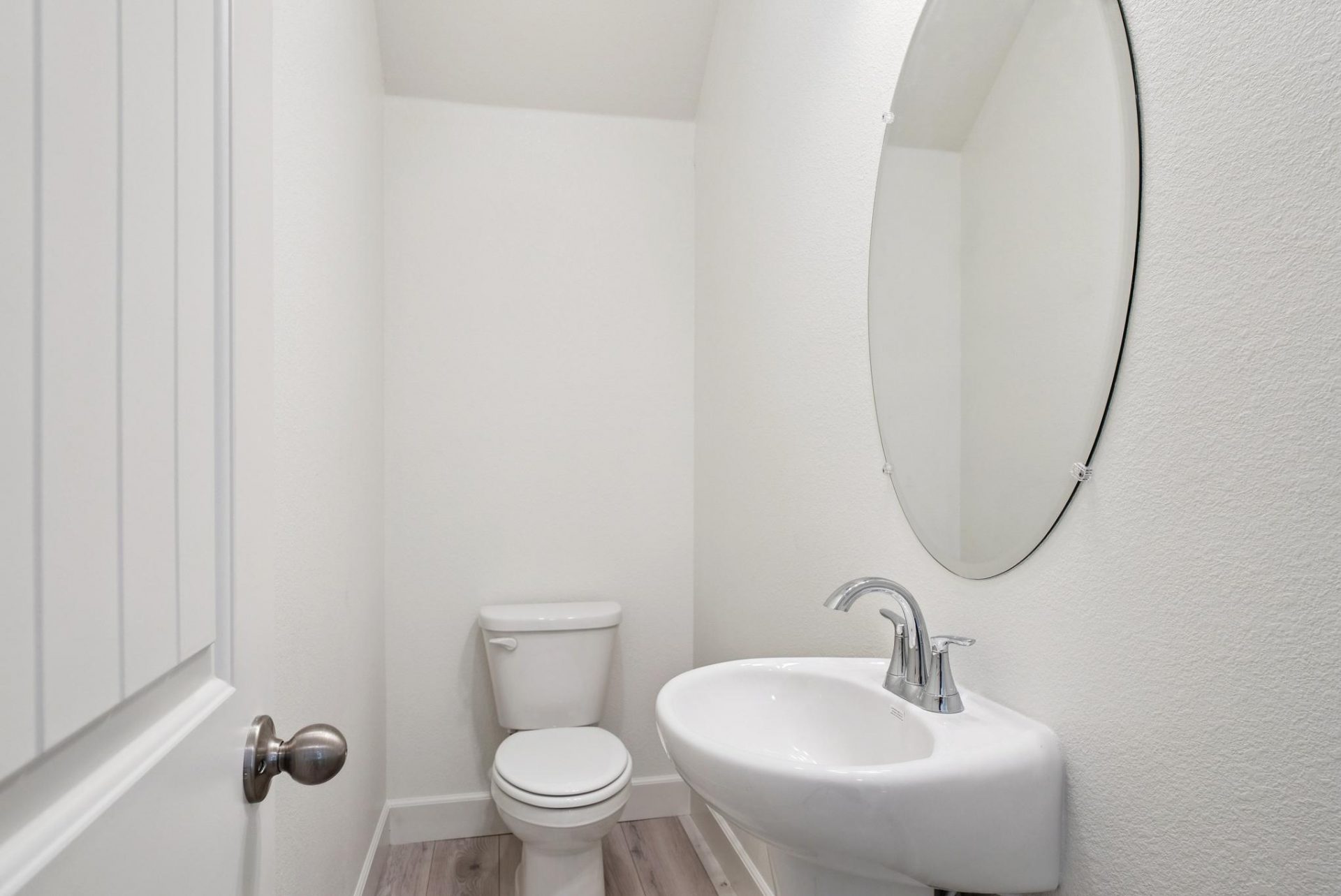 A powder room with a pedestal sink and an oval mirror above it, light colored wood flooring and a toilet.